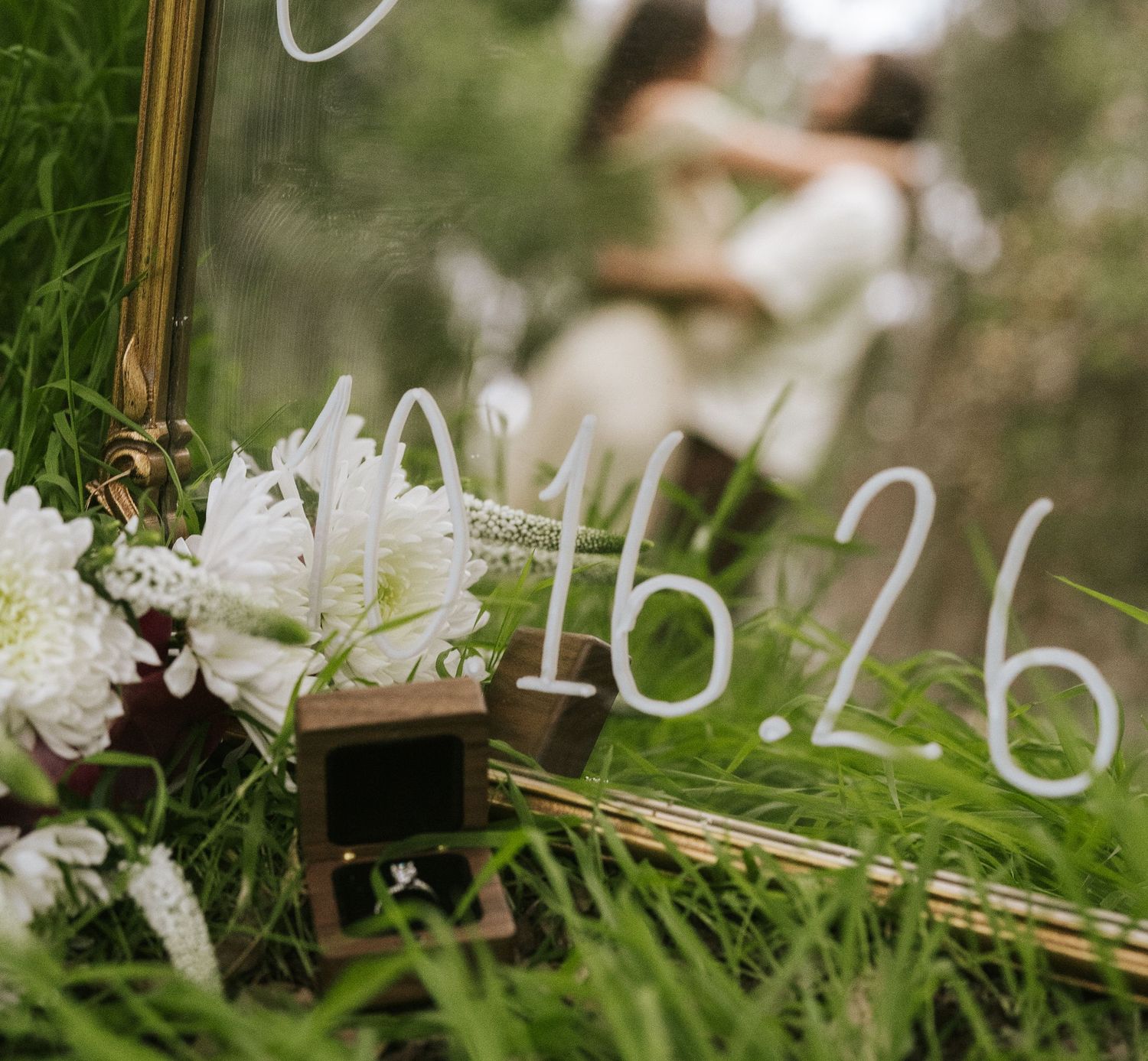 A wedding save-the-date mirror sign with white numbers in a grassy field, flowers, and a blurred couple embracing in the background.