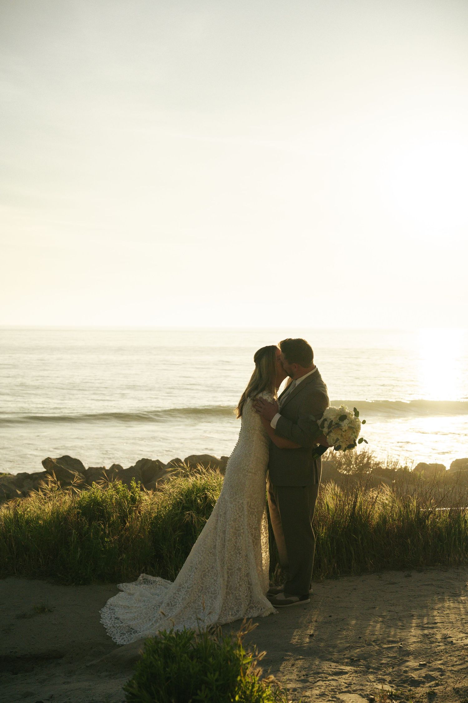 Bride and groom share a romantic kiss at sunset on a rocky beach, bride in lace gown holding bouquet.