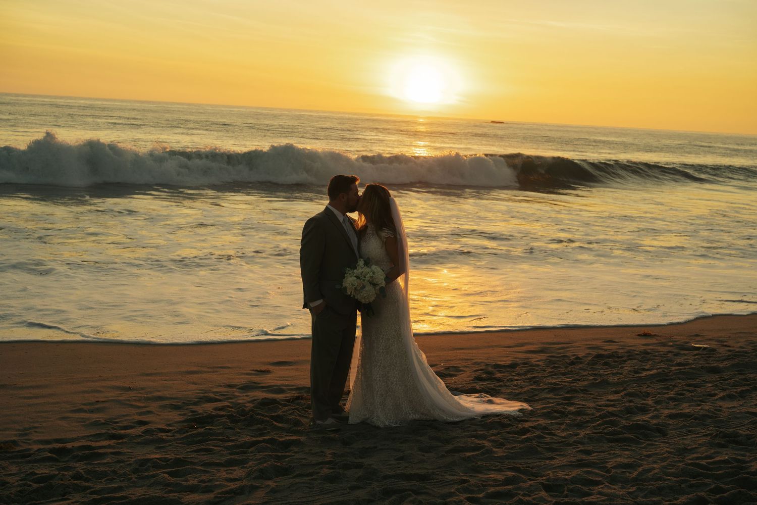Bride and groom share a sunset kiss on the beach with golden waves crashing behind them.
