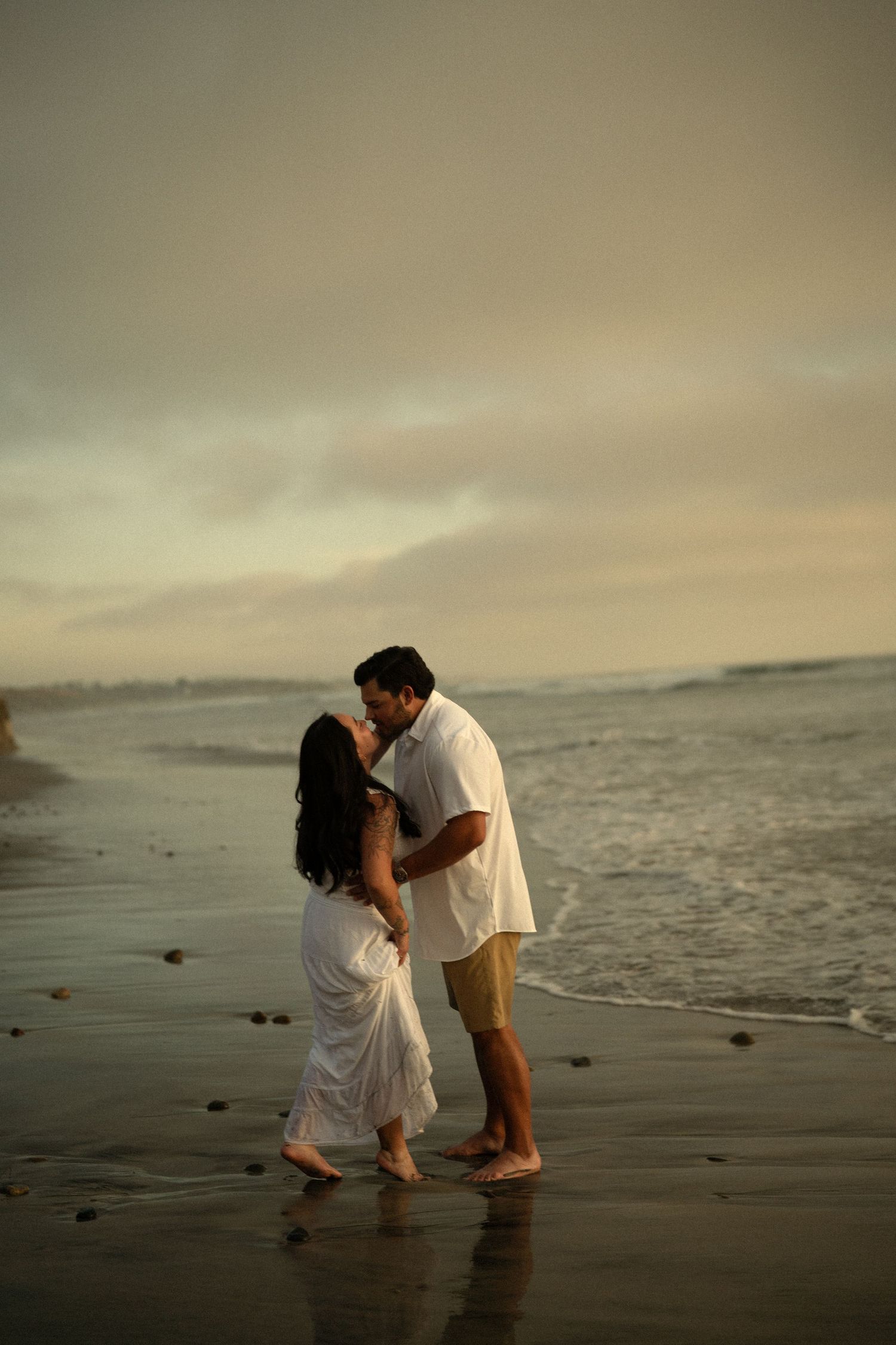 Couple sharing a romantic kiss on a beach at sunset, dressed in white, with ocean waves in the background.