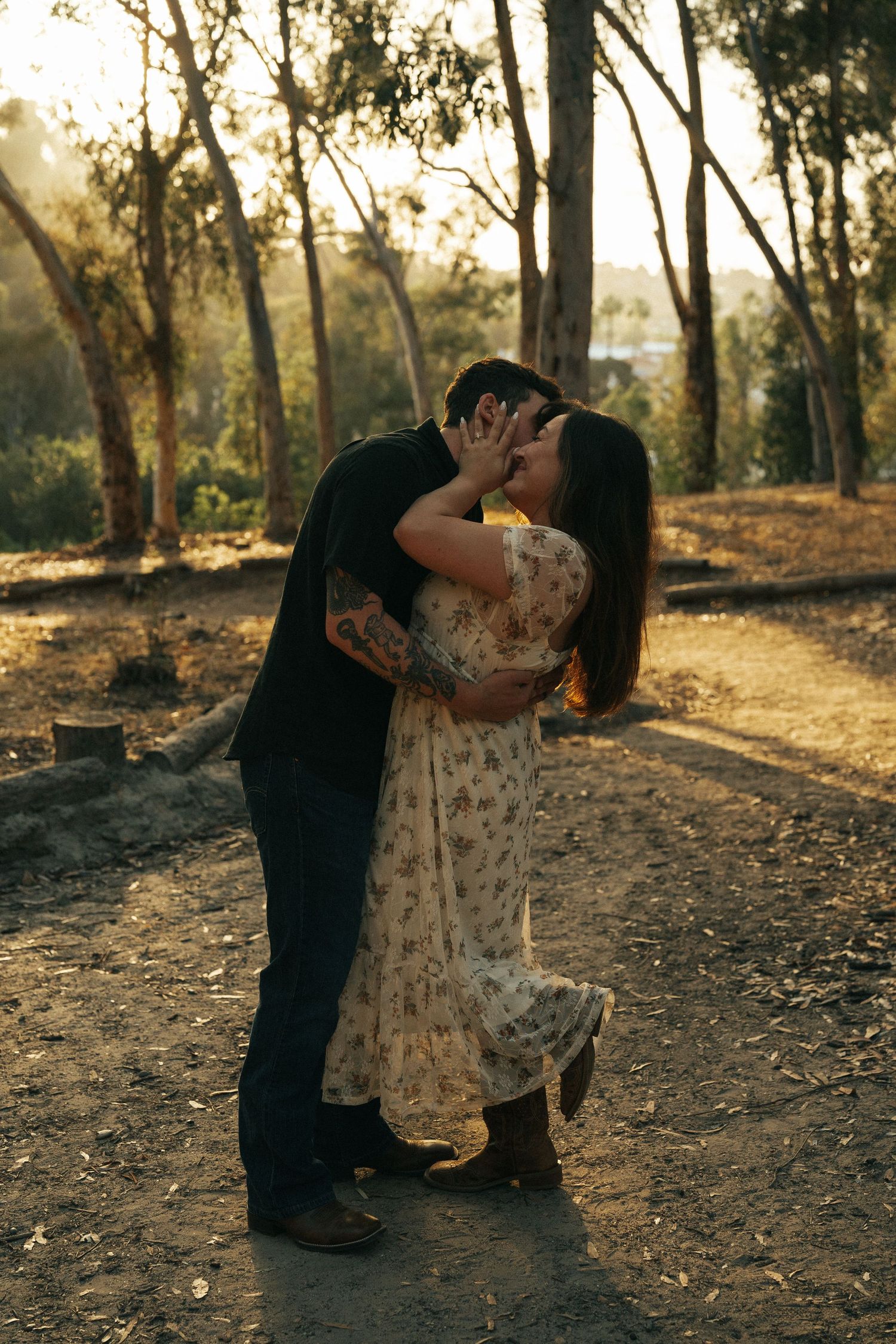 Couple sharing a romantic kiss at golden hour on a forest path, woman in floral dress, man with tattoos.