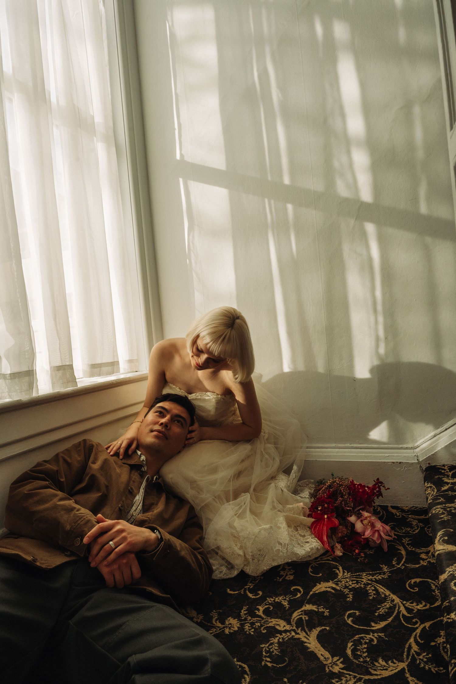 Bride in white gown and groom in brown jacket sit on ornate carpet by sunlit windows with pink bouquet nearby.