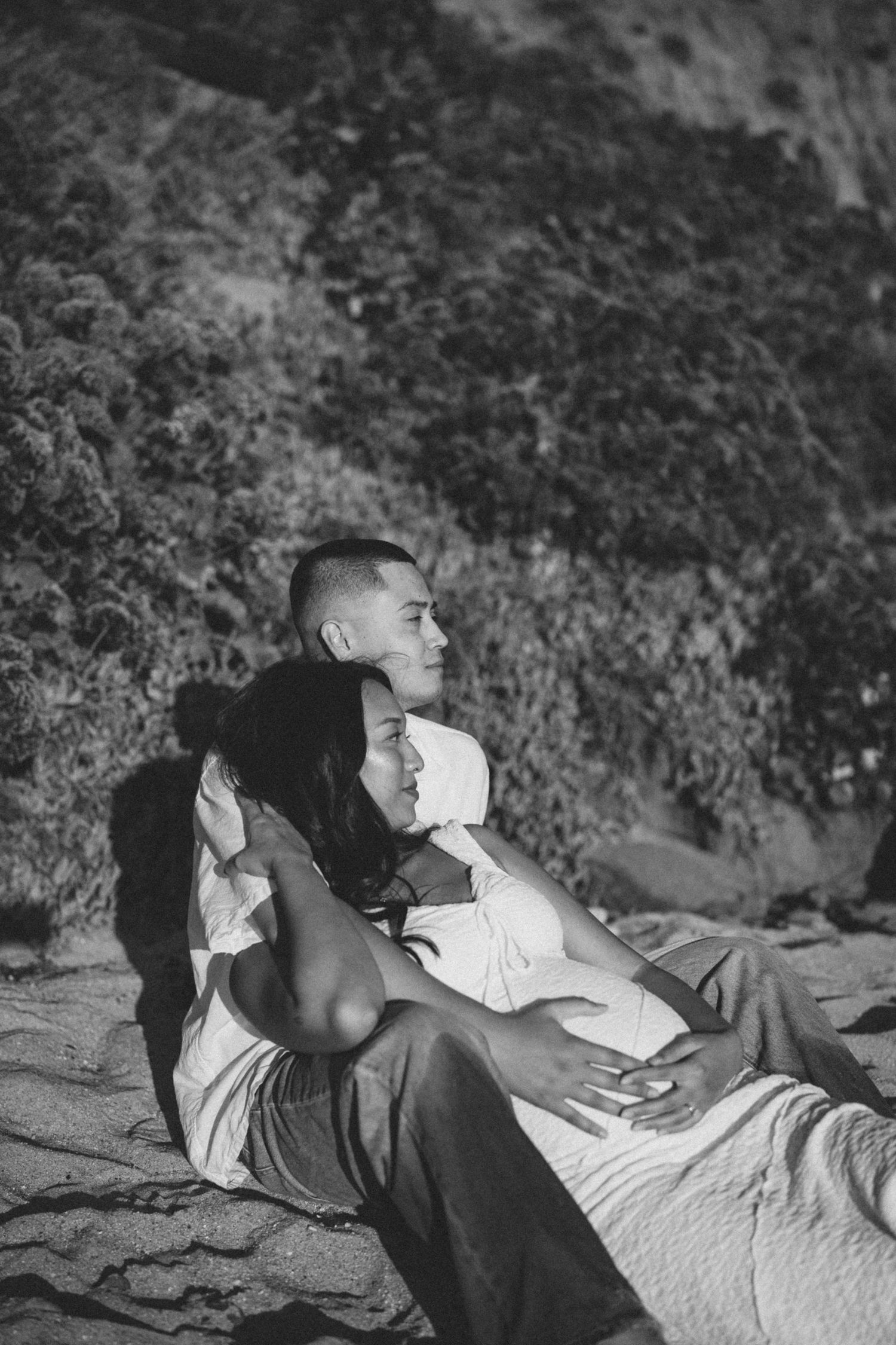 Black and white photo of a couple embracing on sandy beach, sitting together against a rocky hillside backdrop.