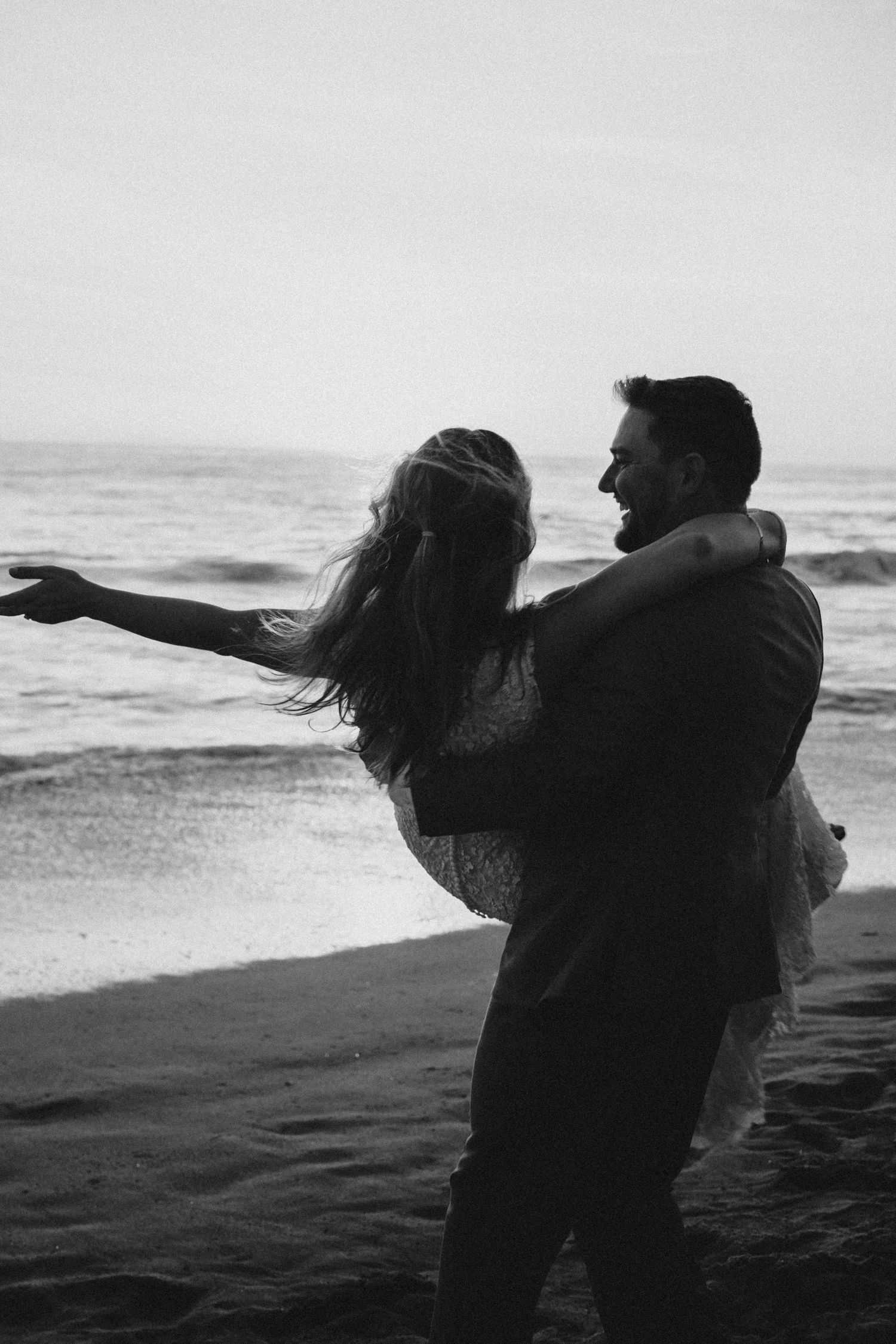 Black and white photo of groom lifting laughing bride on beach with ocean waves behind them.