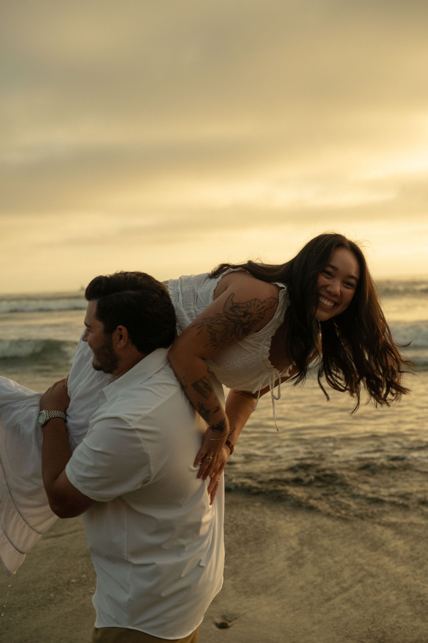 Man giving laughing woman a piggyback ride on the beach at golden sunset, both wearing white.