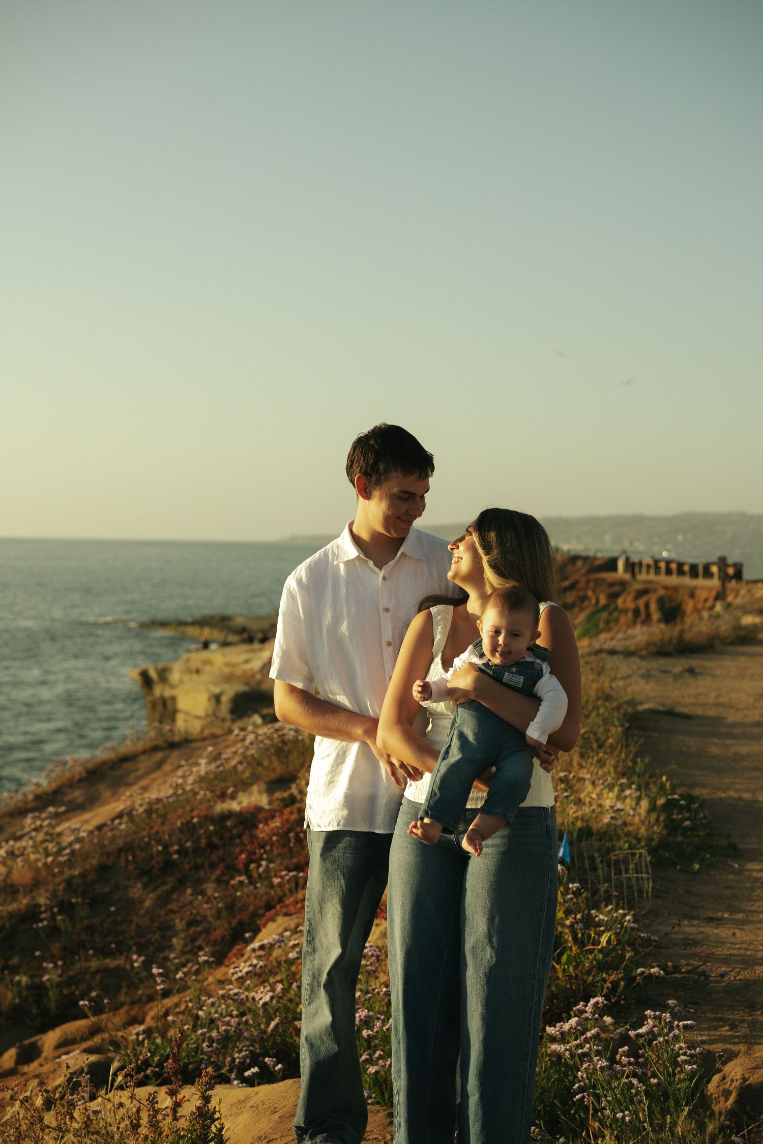 Young family with baby enjoying golden hour at rocky ocean cliffside, parents gazing lovingly at infant.
