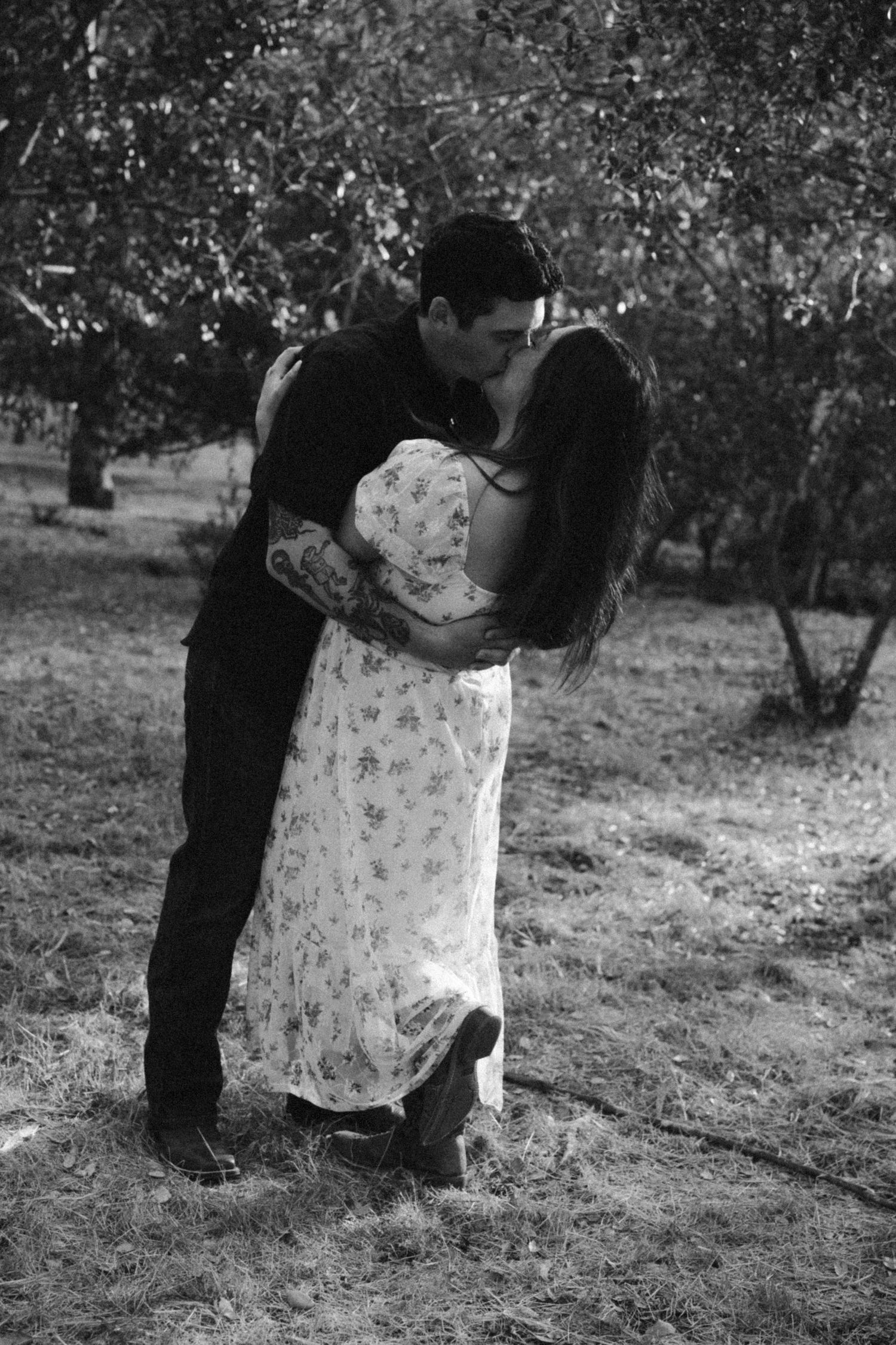 Black and white photo of a couple sharing a passionate kiss in an outdoor orchard setting.