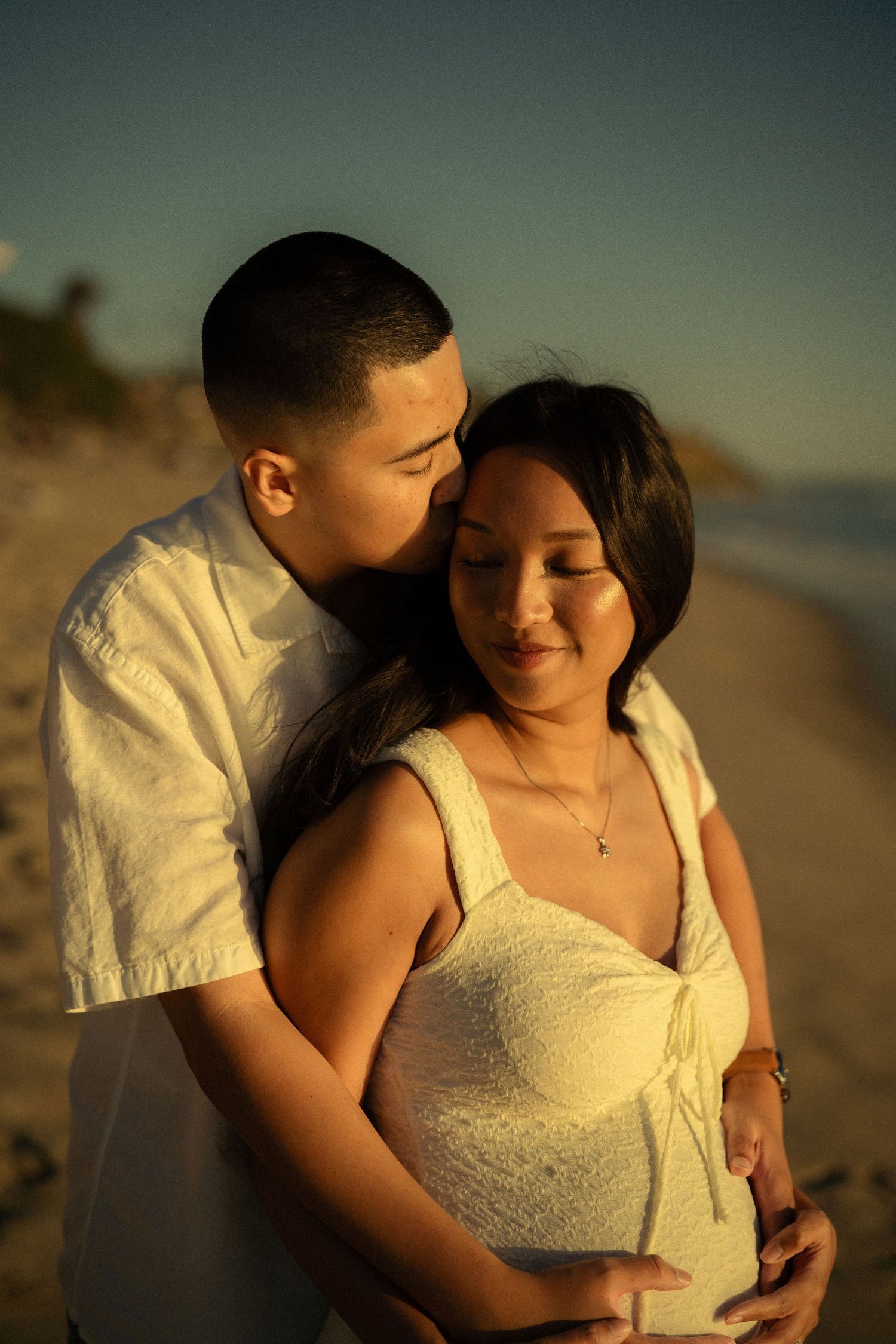 Couple sharing tender moment on beach at golden hour, man kissing pregnant woman's cheek while cradling her belly.