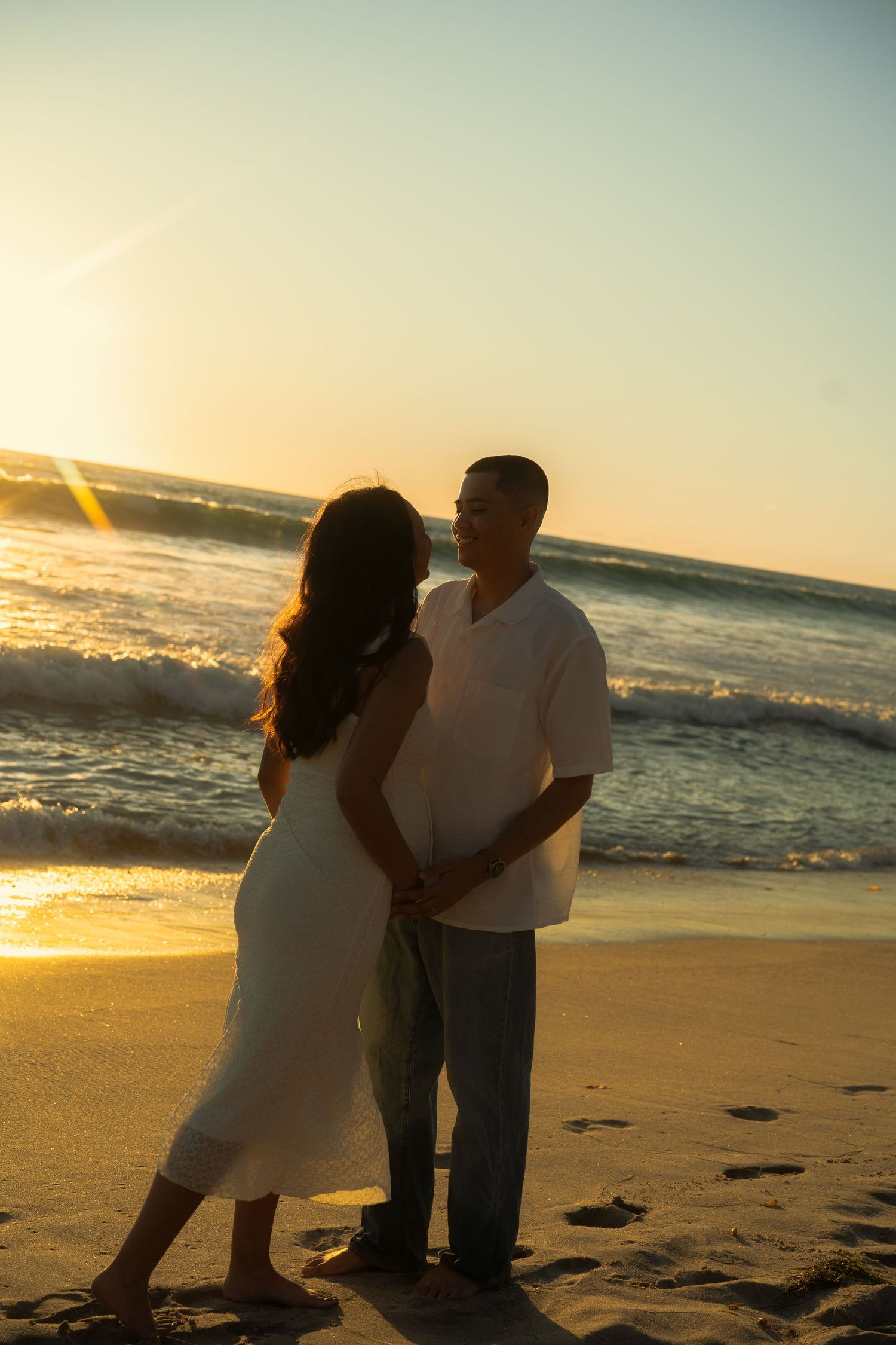 Couple holding hands standing together on sandy beach during warm golden sunset.