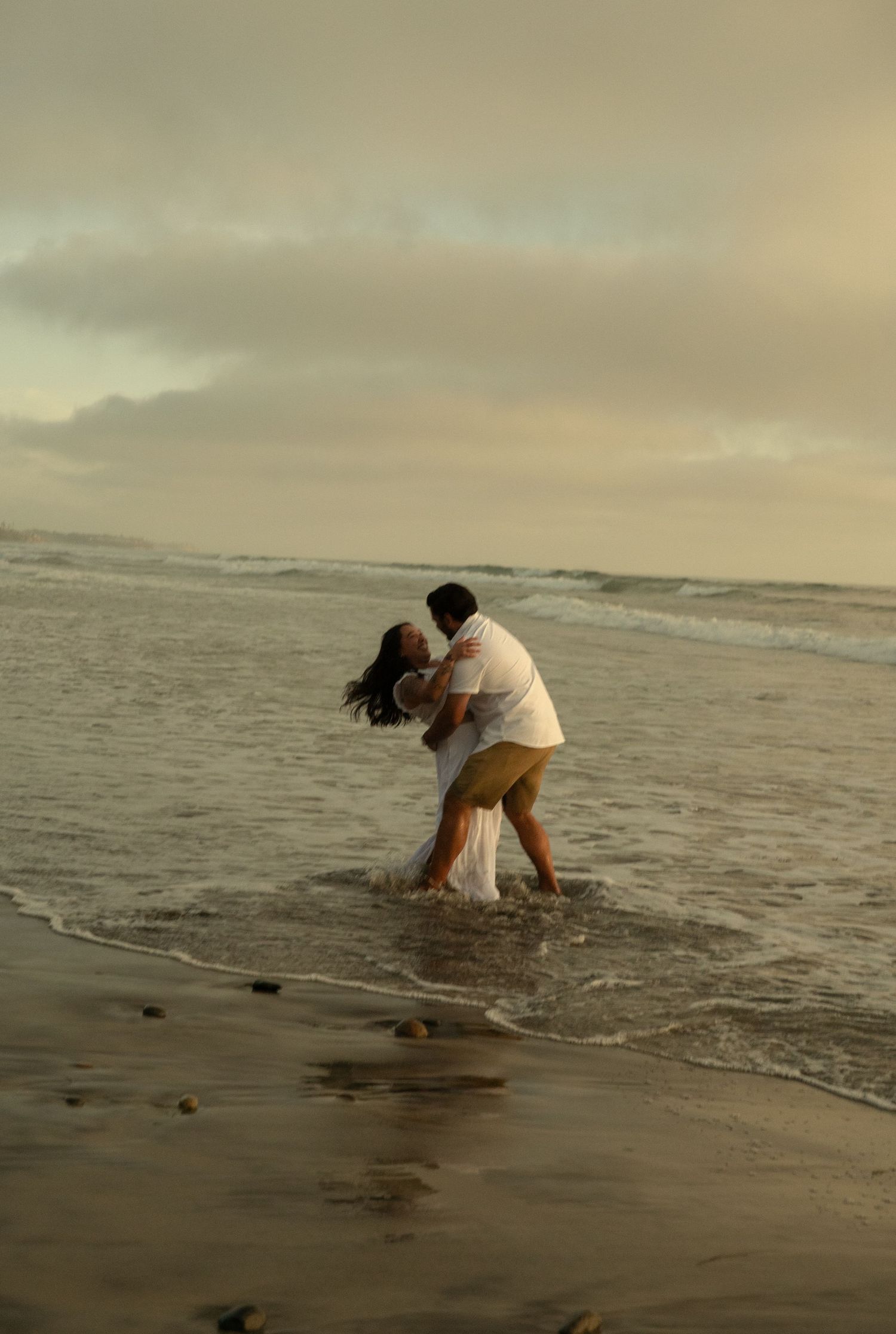 Couple embracing and kissing in ocean waves on the beach during a dramatic cloudy sunset.