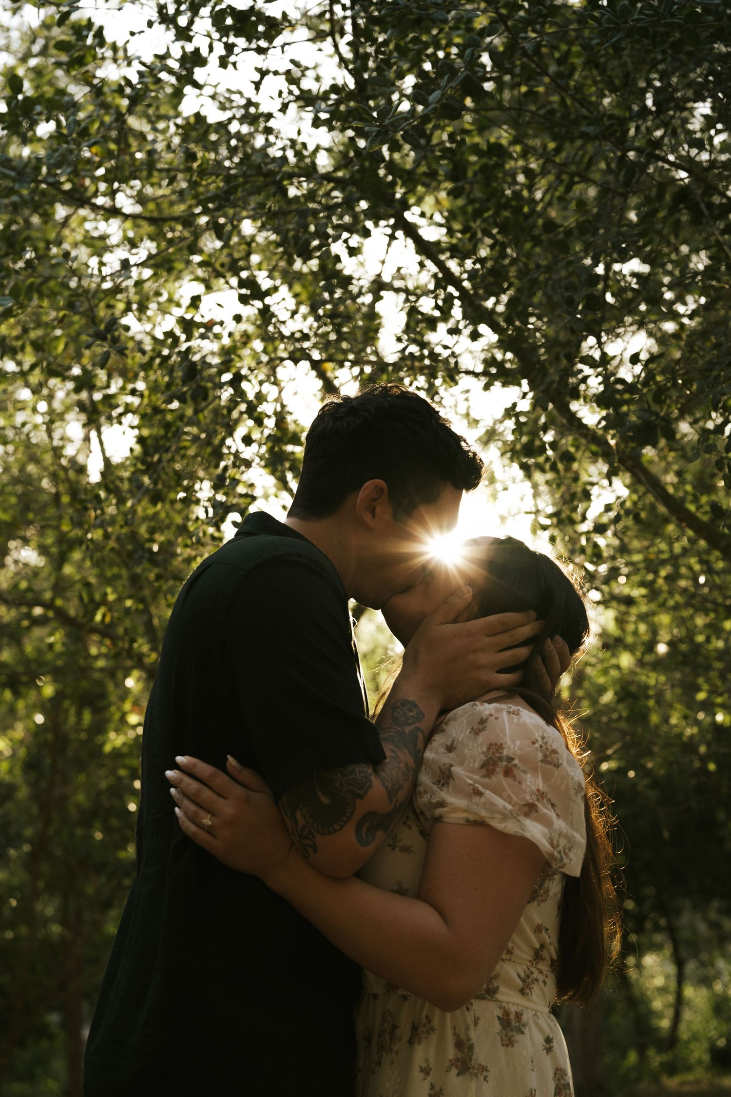Couple sharing a romantic kiss in a sunlit garden, with golden light streaming through lush green trees behind them.
