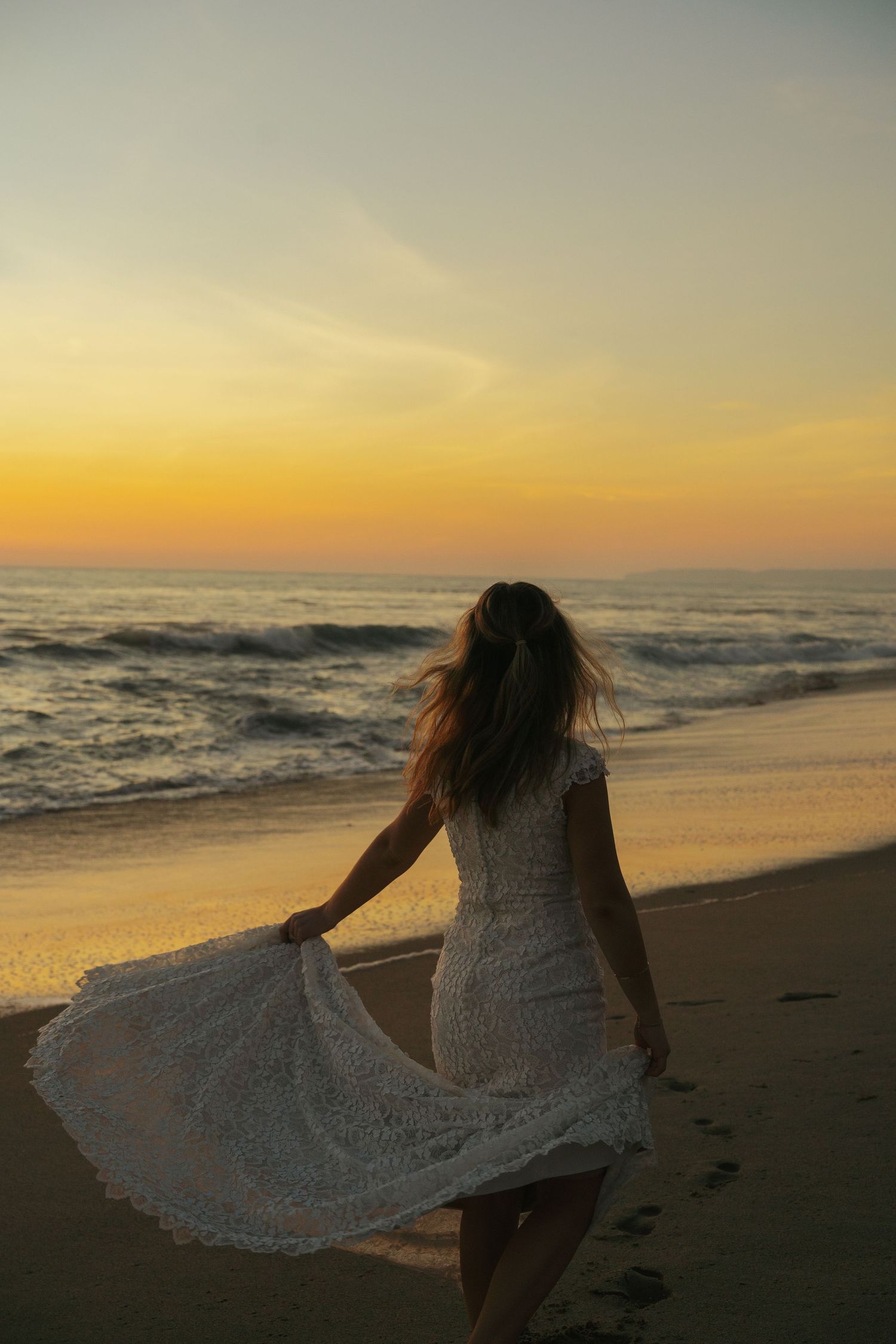 Bride in white lace dress walks along sandy beach at sunset, dress flowing in the ocean breeze.