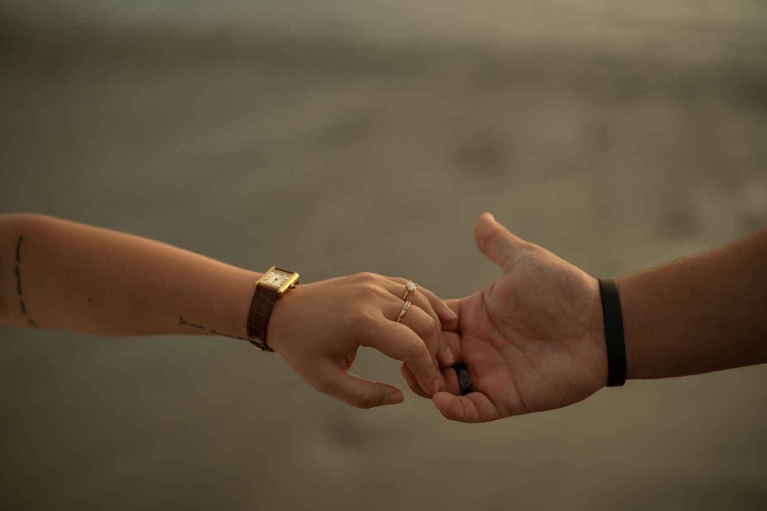 Two hands gently touching fingers, one wearing a gold watch and ring, the other a black bracelet, against a blurred background.