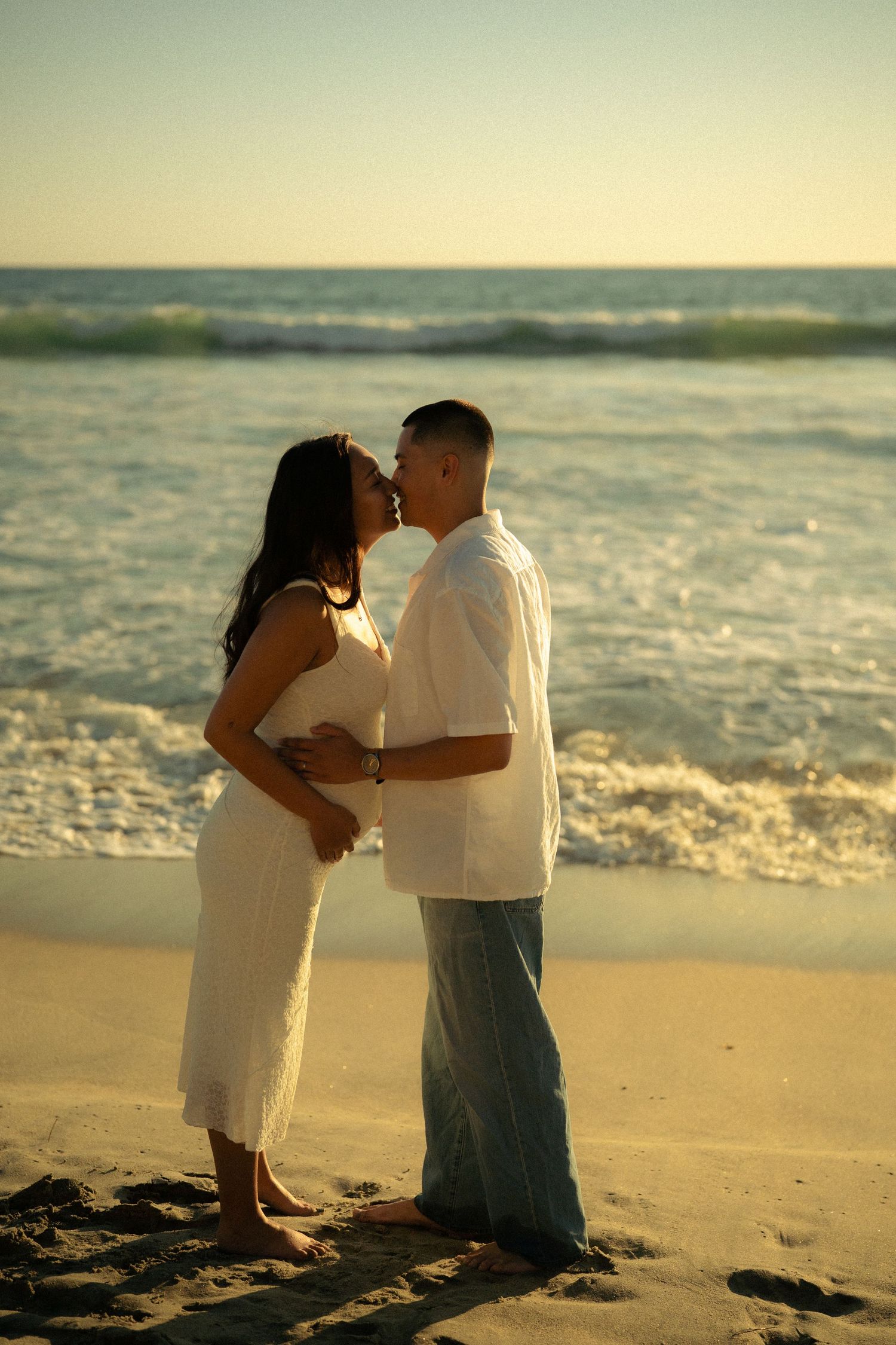 Pregnant couple sharing a tender kiss on a sandy beach at sunset, waves crashing behind them.