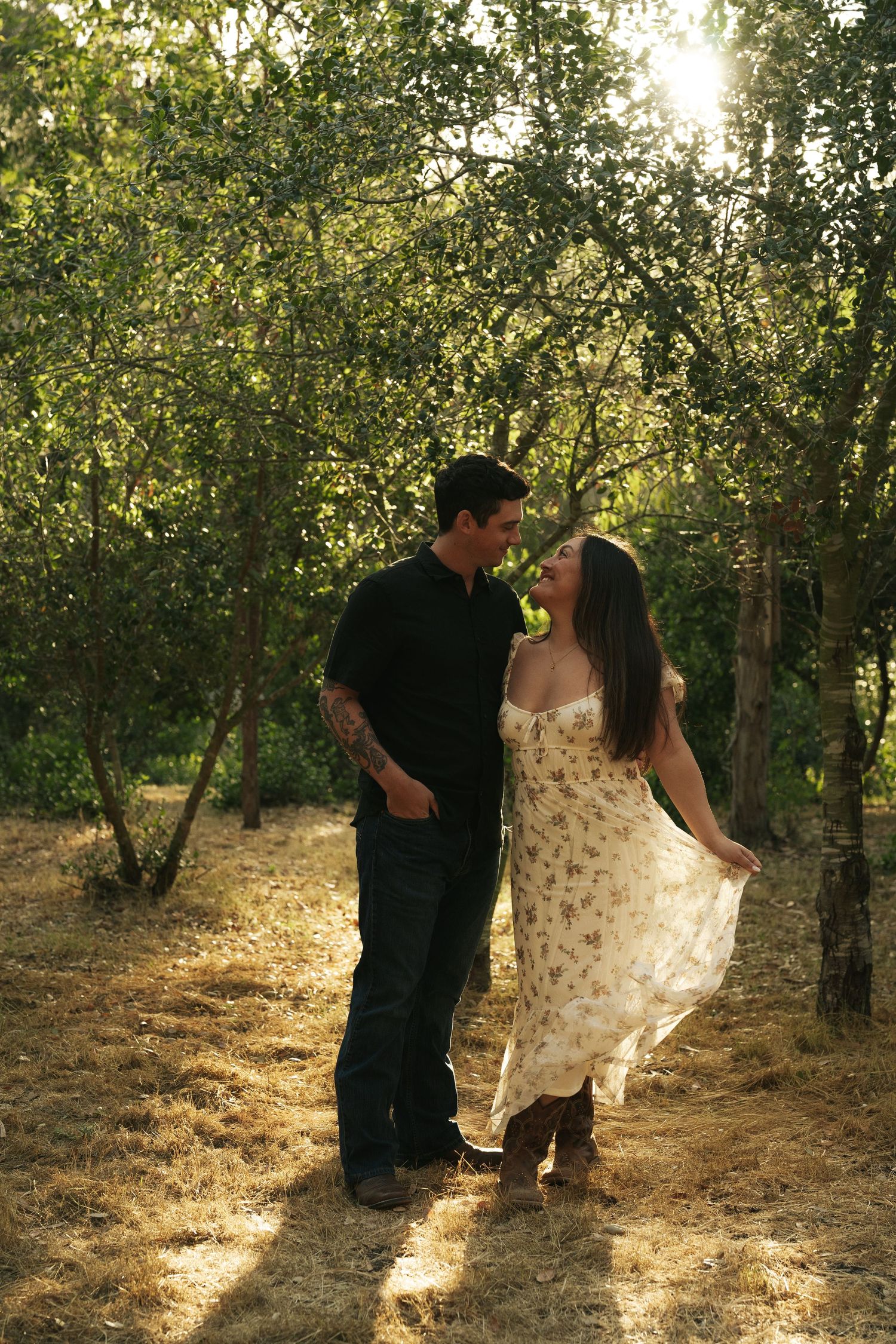 A couple shares a romantic moment in a sunlit orchard, woman in floral dress holding her skirt gently.