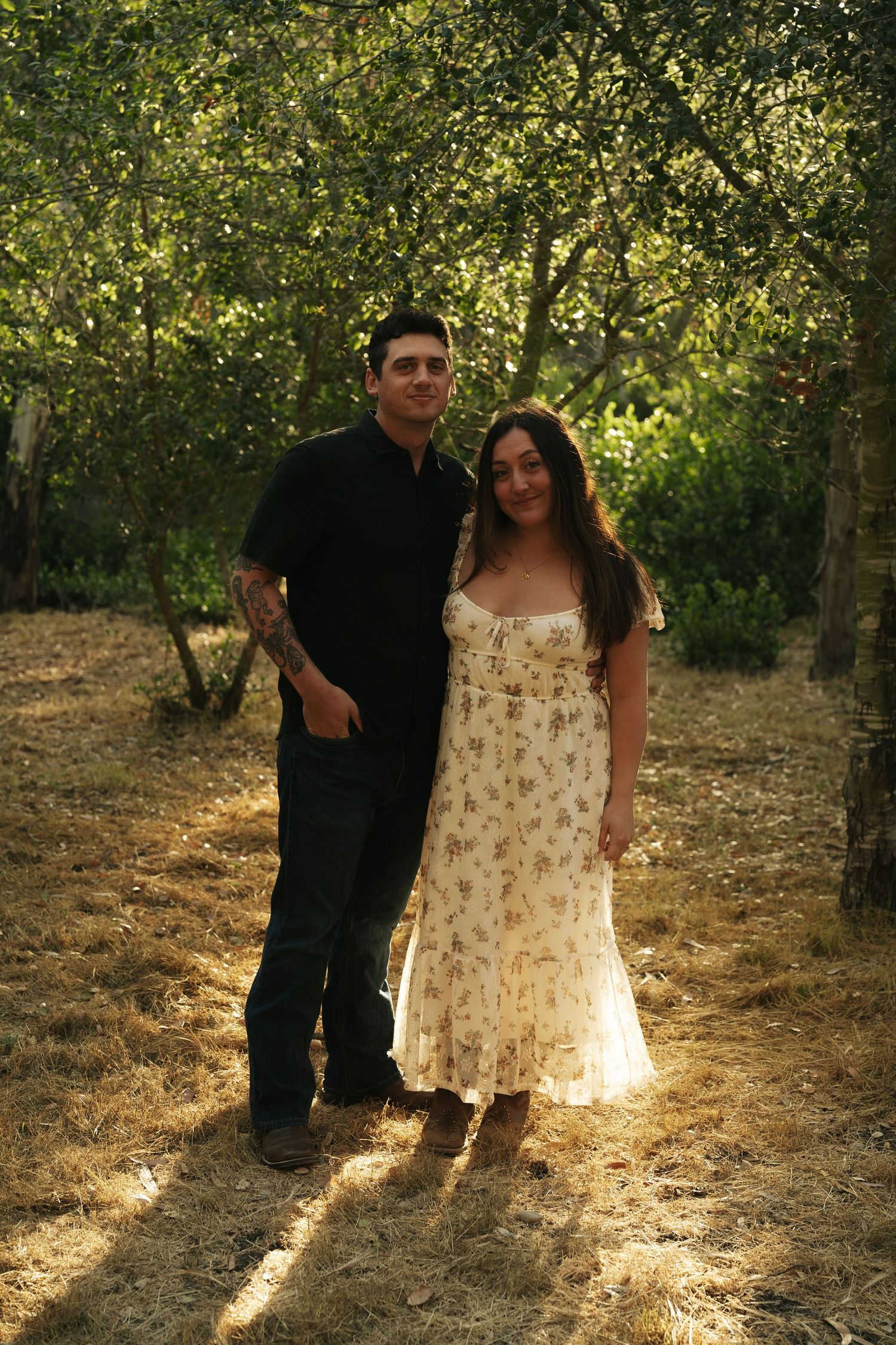 A couple poses together in a sunlit orchard, woman in floral dress and man in black shirt smiling at camera.