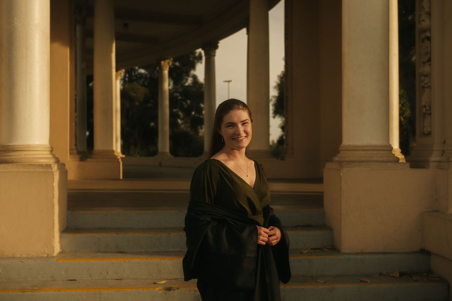 A smiling woman in a dark outfit stands between elegant columns at golden hour.