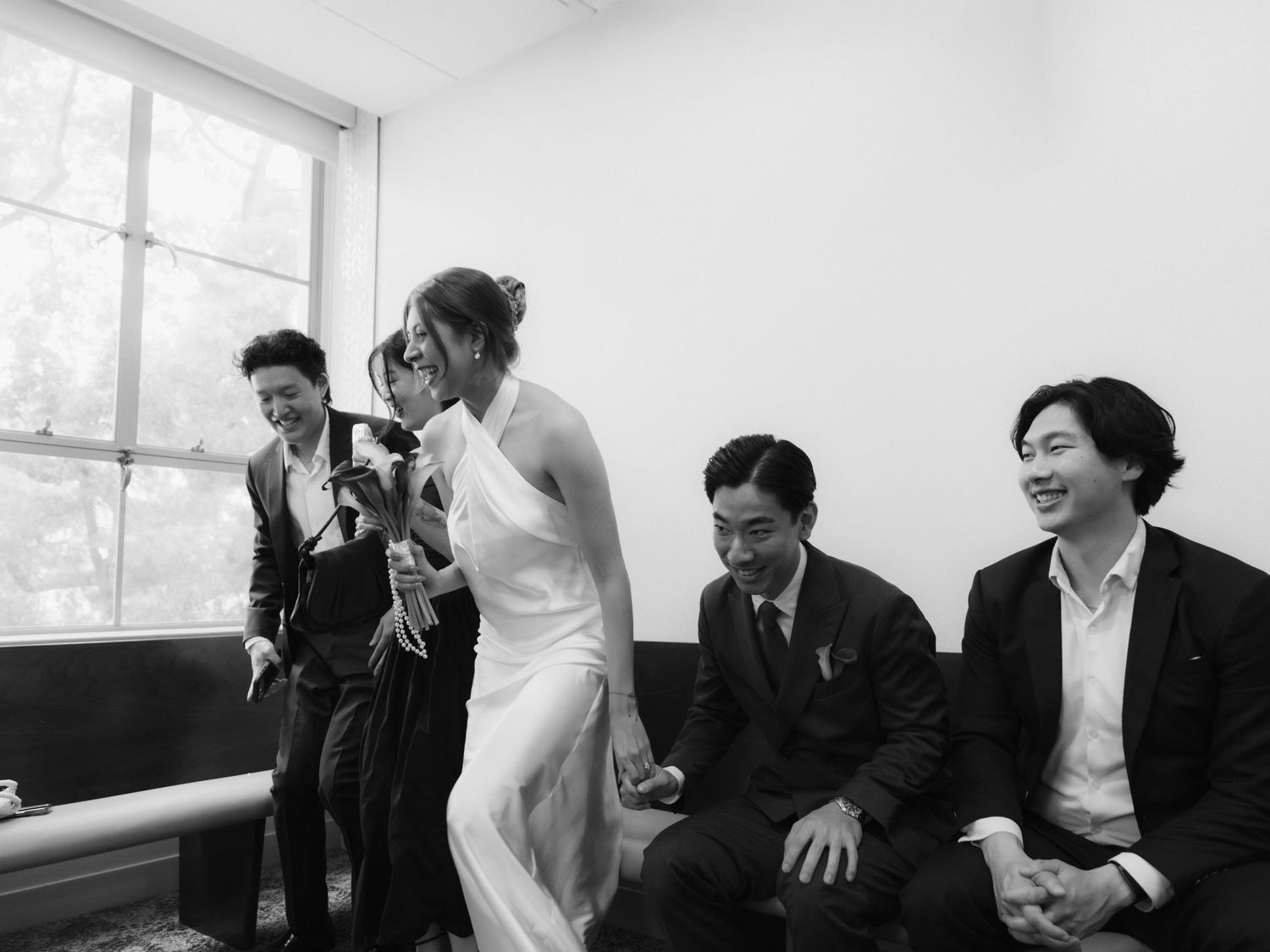 Black and white wedding photo of a bride in white dress holding flowers with groom and guests seated nearby.