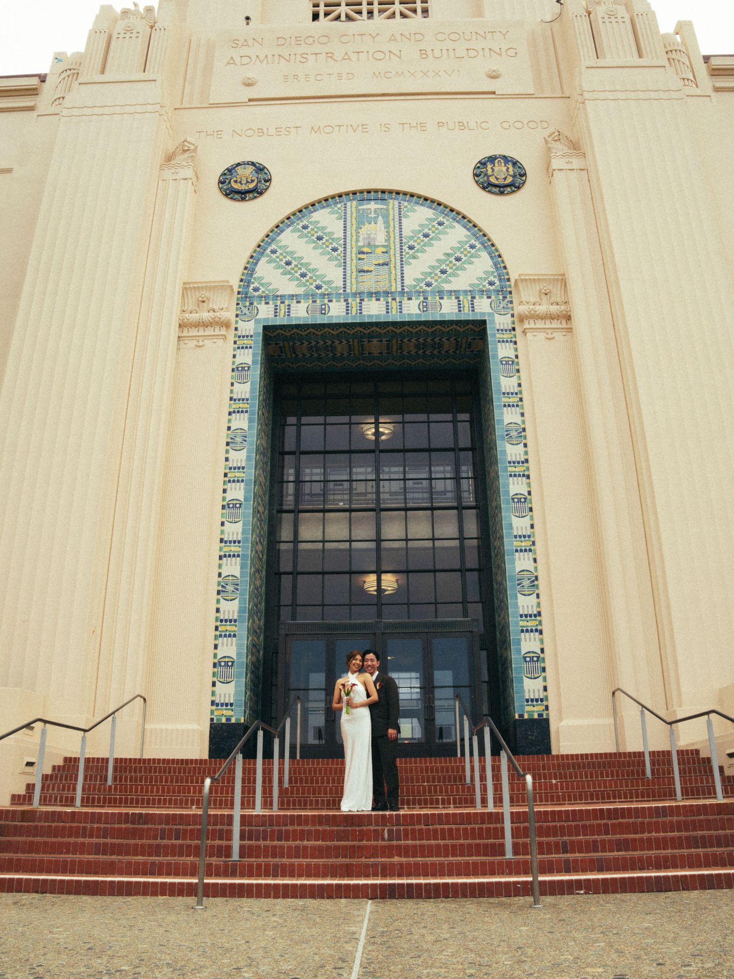 Bride and groom posing on steps of ornate Art Deco building with decorative arch and tiled facade.