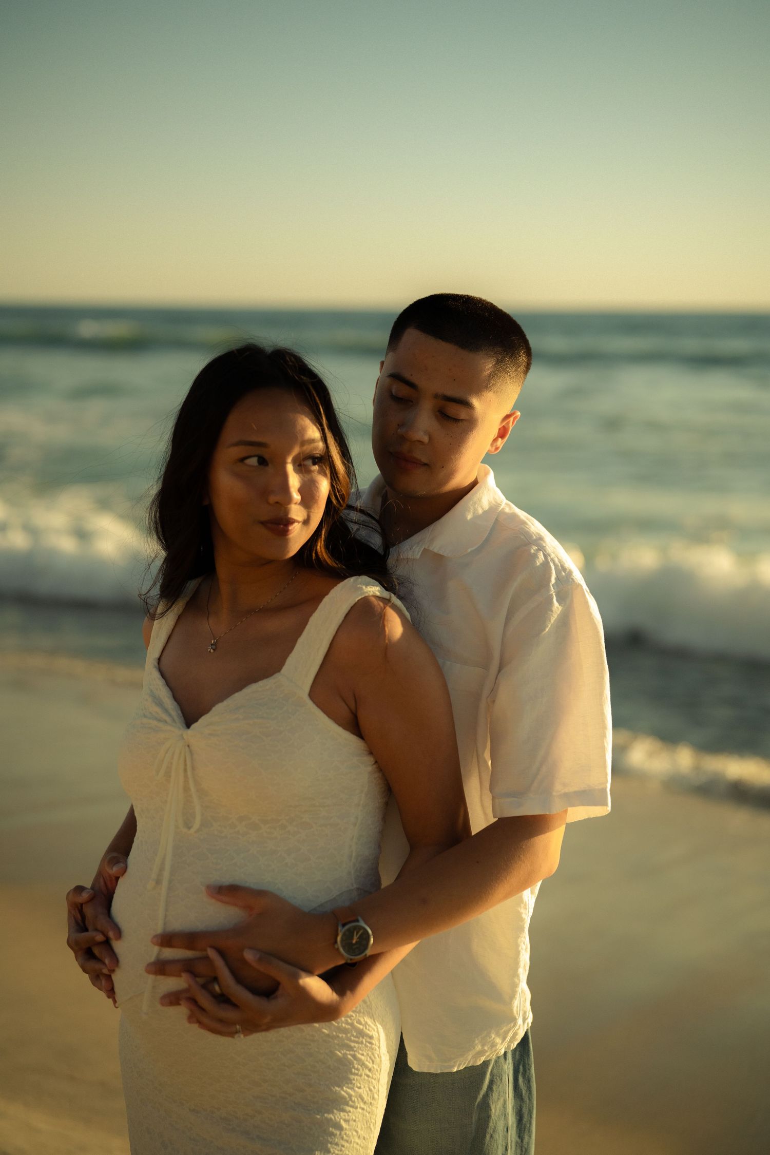 Expecting couple embracing on beach at golden hour, man holding pregnant woman's belly with ocean backdrop.