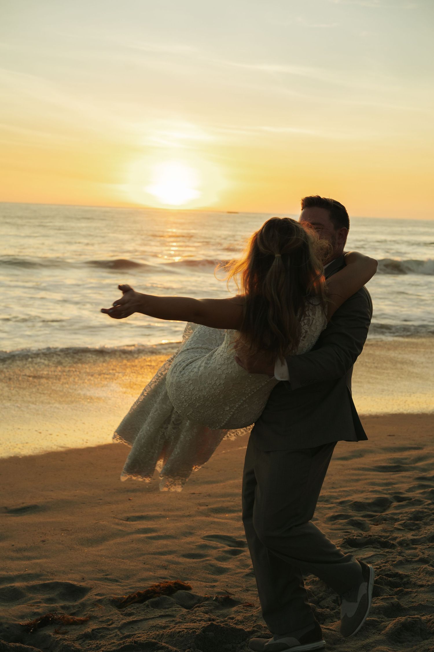 Groom lifts bride in white lace dress on sandy beach during vibrant golden sunset over ocean waves.