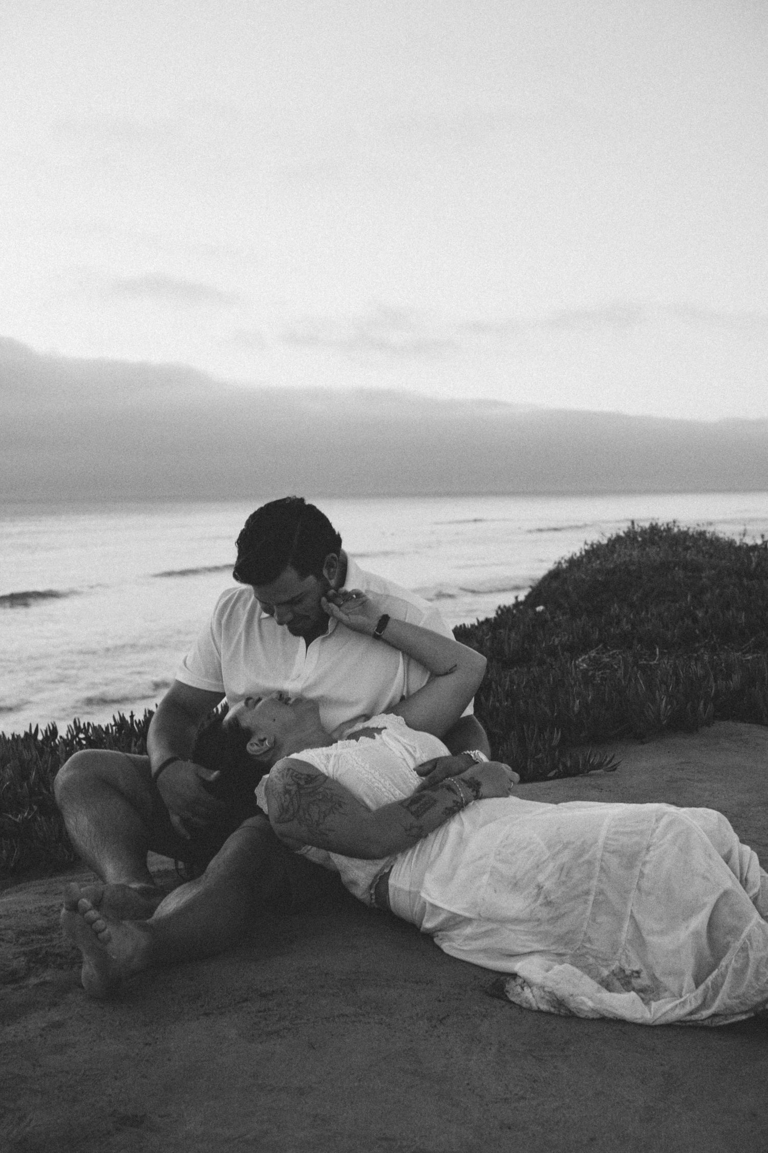 Black and white photo of couple in romantic pose on coastal cliffside, ocean waves in background.