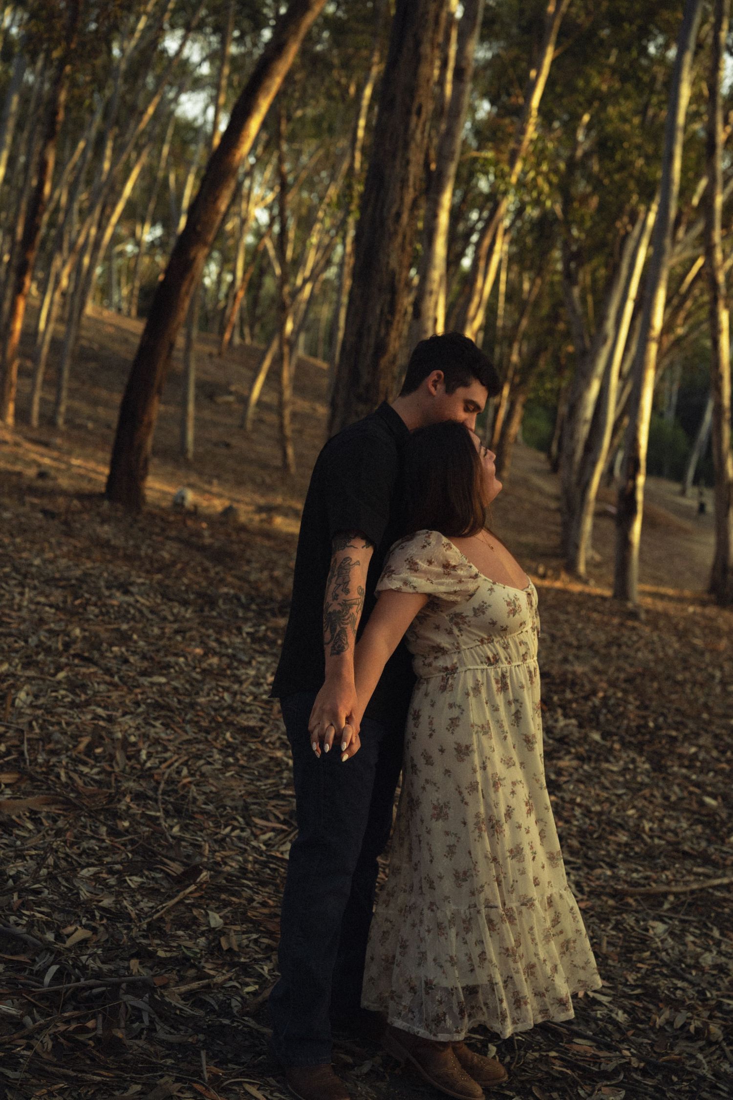 A couple shares a romantic moment holding hands in a sunlit forest, the woman wearing a floral dress.