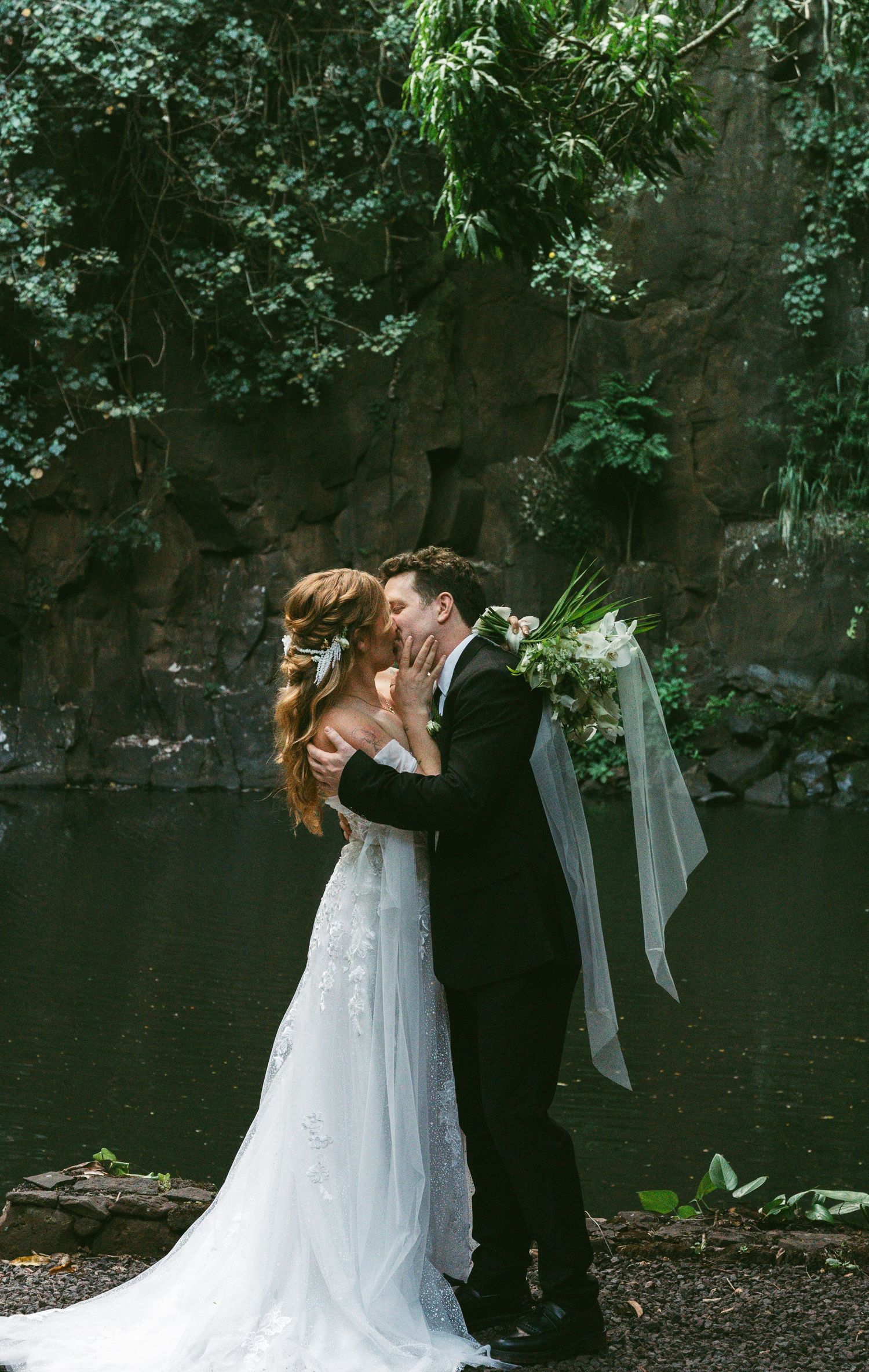 Bride and groom share a romantic kiss in lush garden setting, bride in lace gown holding white bouquet.