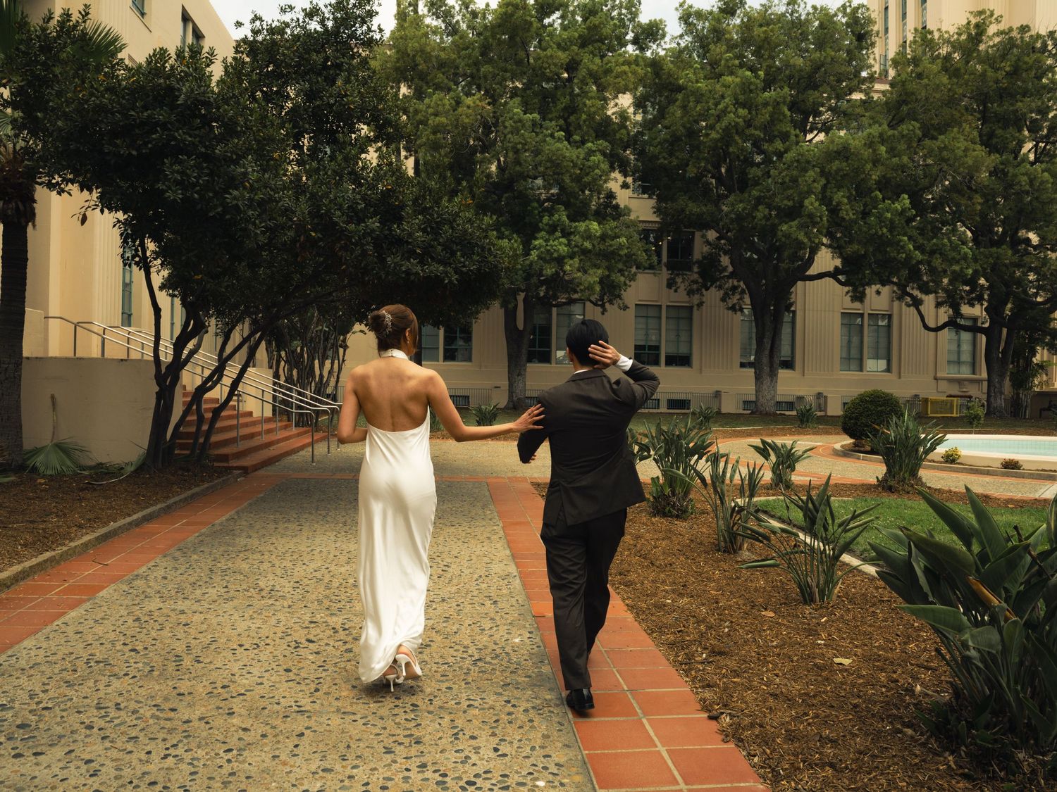 Bride and groom walking hand in hand down a garden pathway, bride in backless white gown, groom in black suit.