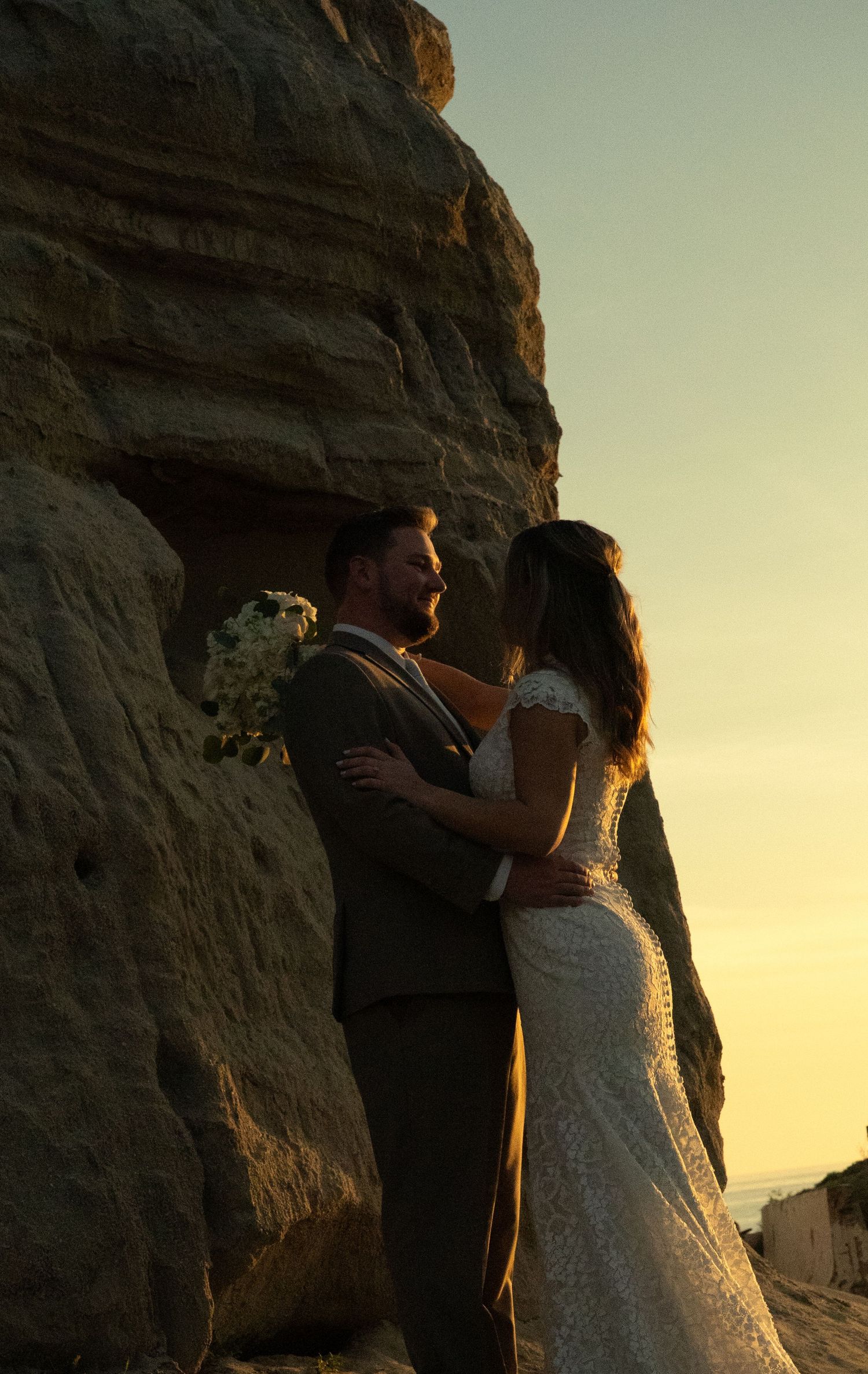 Bride and groom embrace romantically beside dramatic sandstone cliffs at golden sunset.