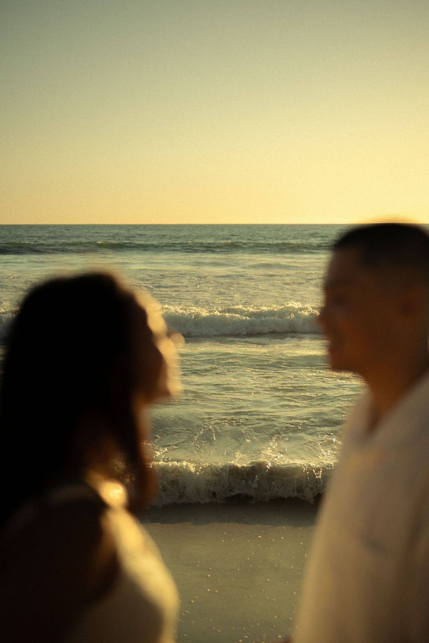 Blurred couple standing together on beach at golden sunset, gazing at ocean waves in soft focus.