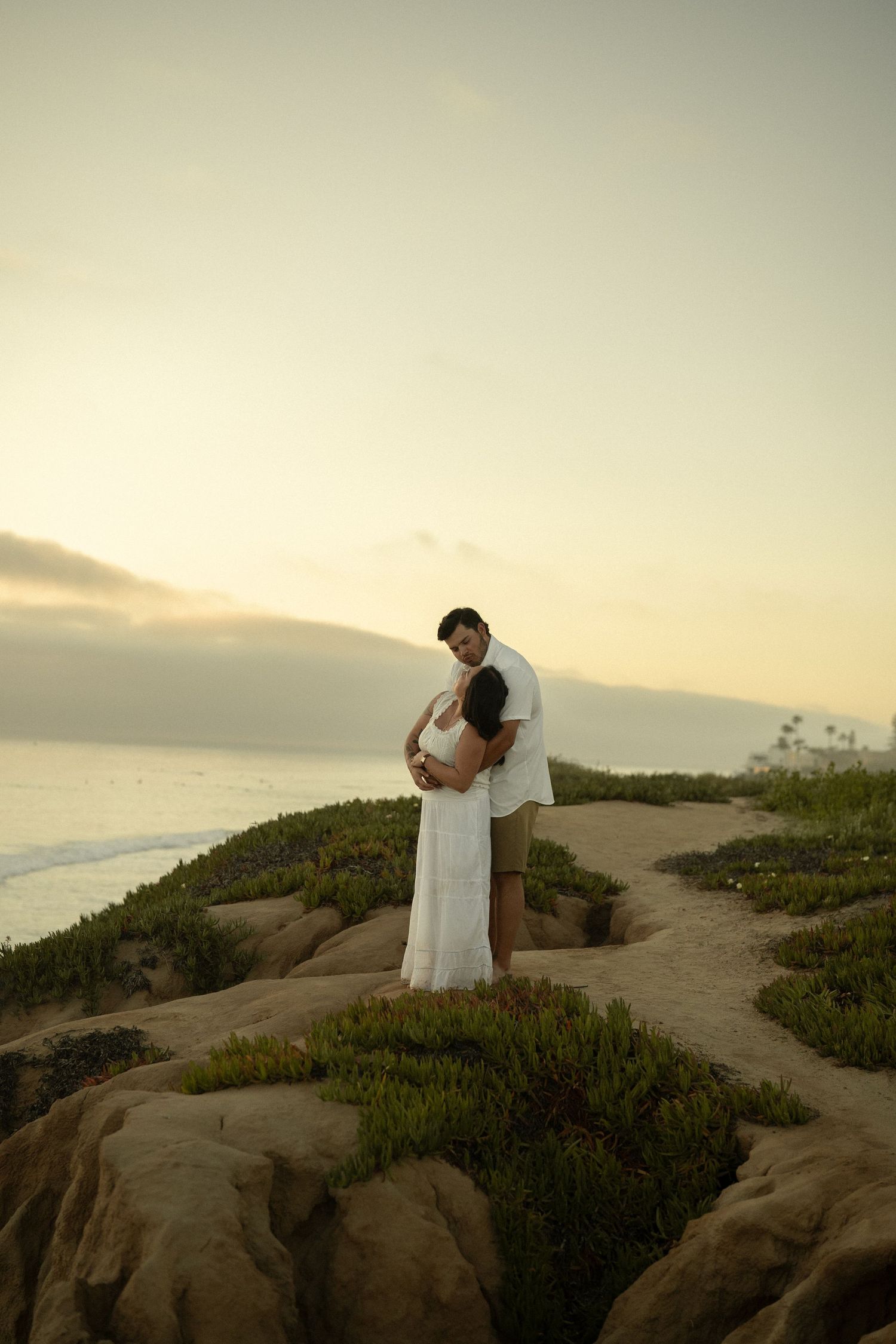 Couple sharing tender embrace on scenic coastal cliffs at sunset, woman in flowing white dress, ocean in background.