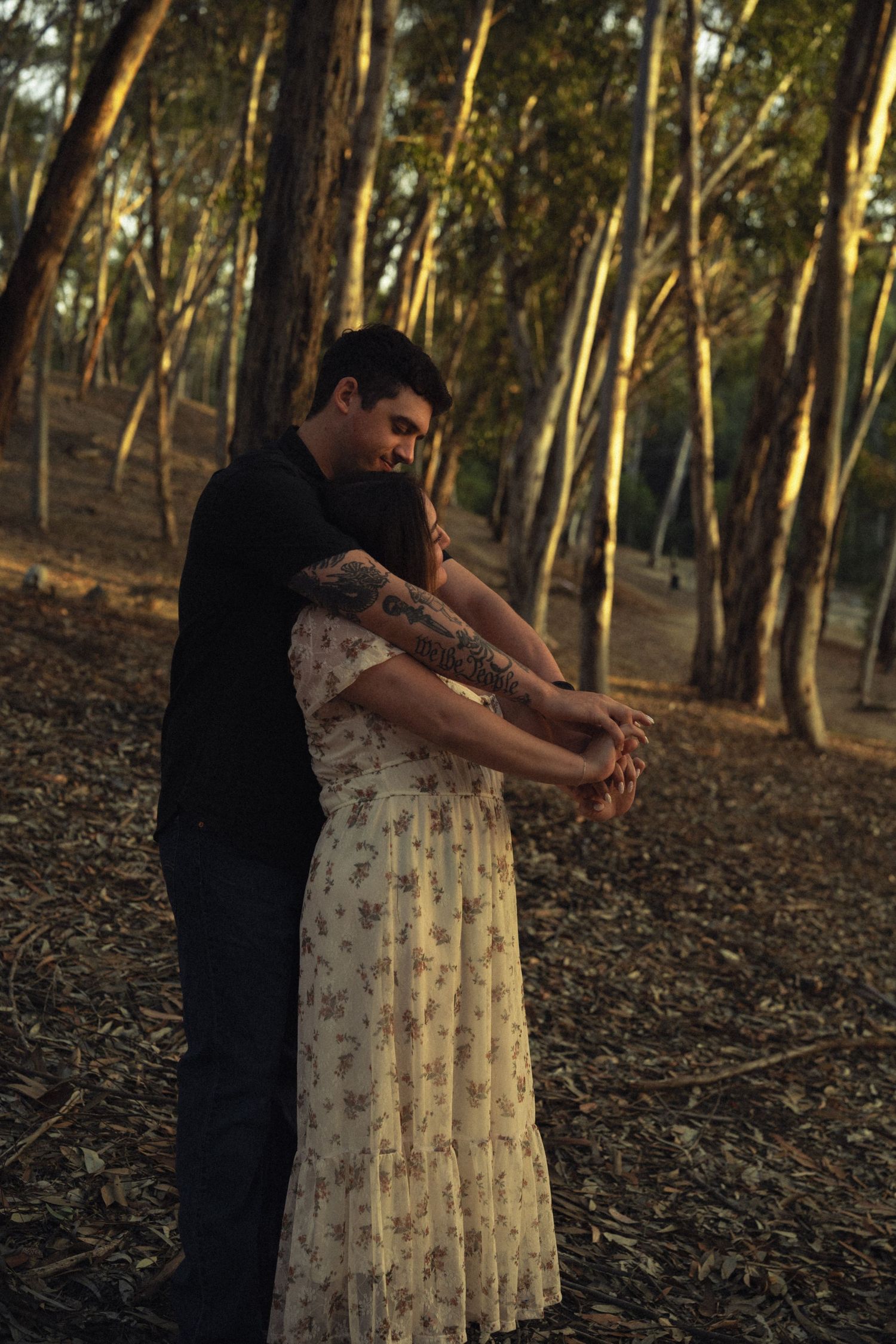 Man embracing woman from behind while holding hands in a warm sunlit woodland setting.