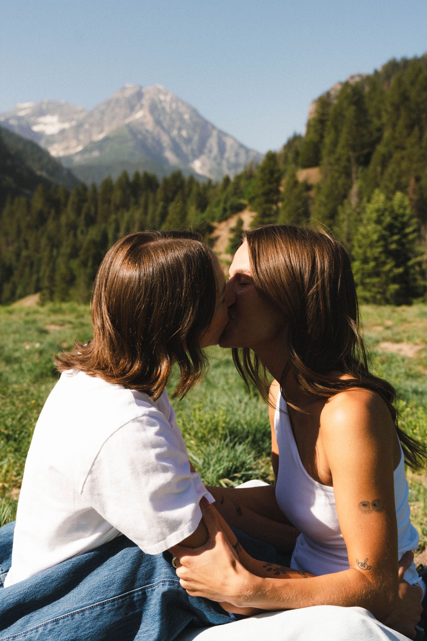 Two people embrace while sitting in an alpine meadow with mountain peaks in the distance.