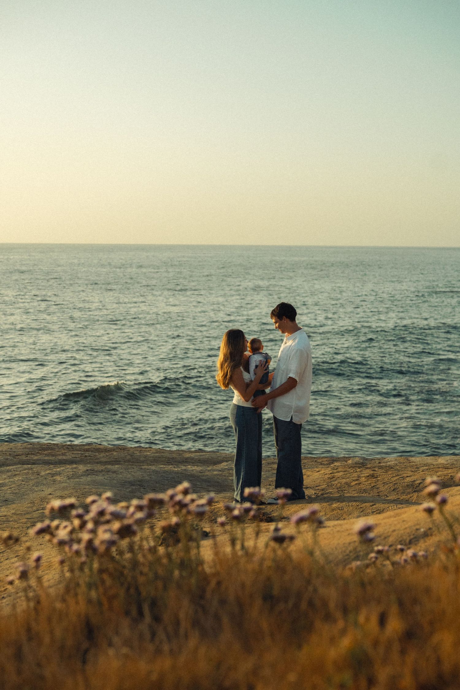 Young family standing together on coastal cliffs with wildflowers and ocean view at sunset.