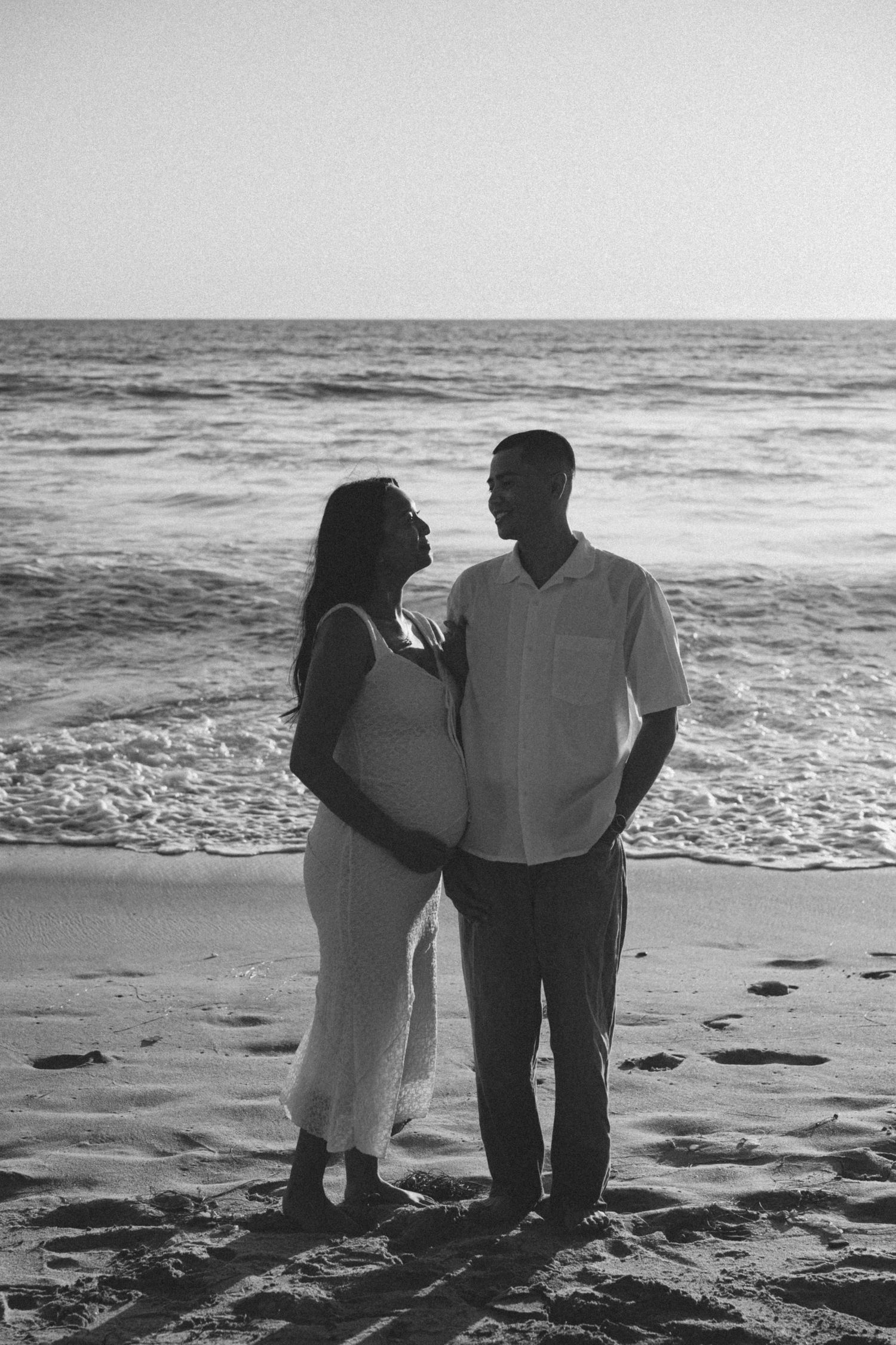 Black and white photo of pregnant couple standing together on beach, ocean waves behind them.