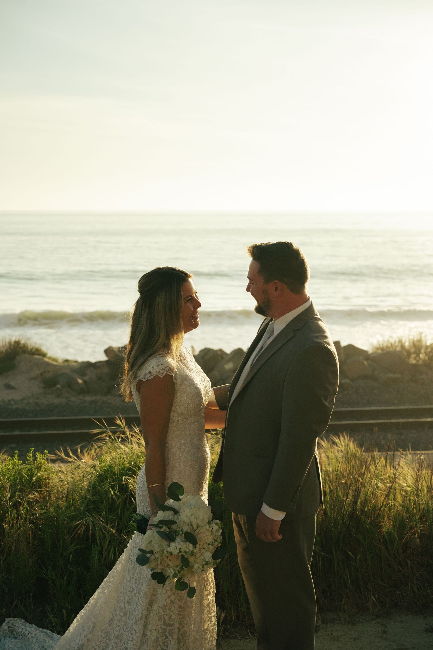Bride and groom share a romantic moment on a beach at sunset, with ocean waves in the background.