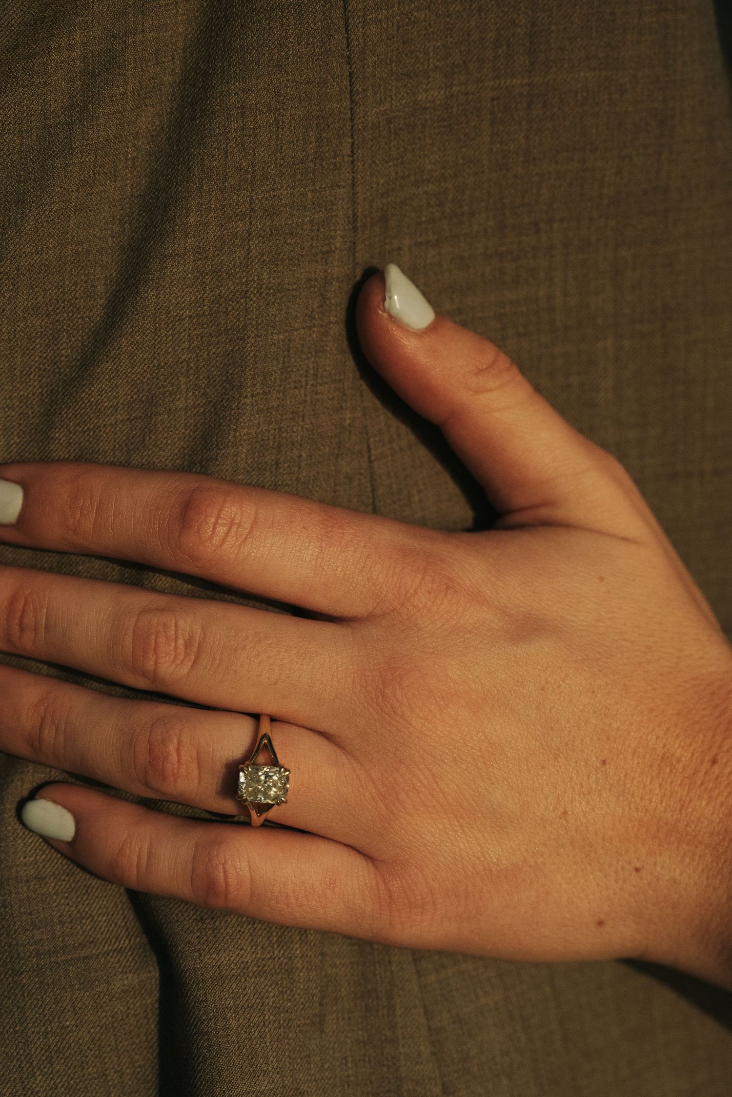Close-up of a hand with light blue nails wearing a gold solitaire diamond engagement ring against olive fabric.
