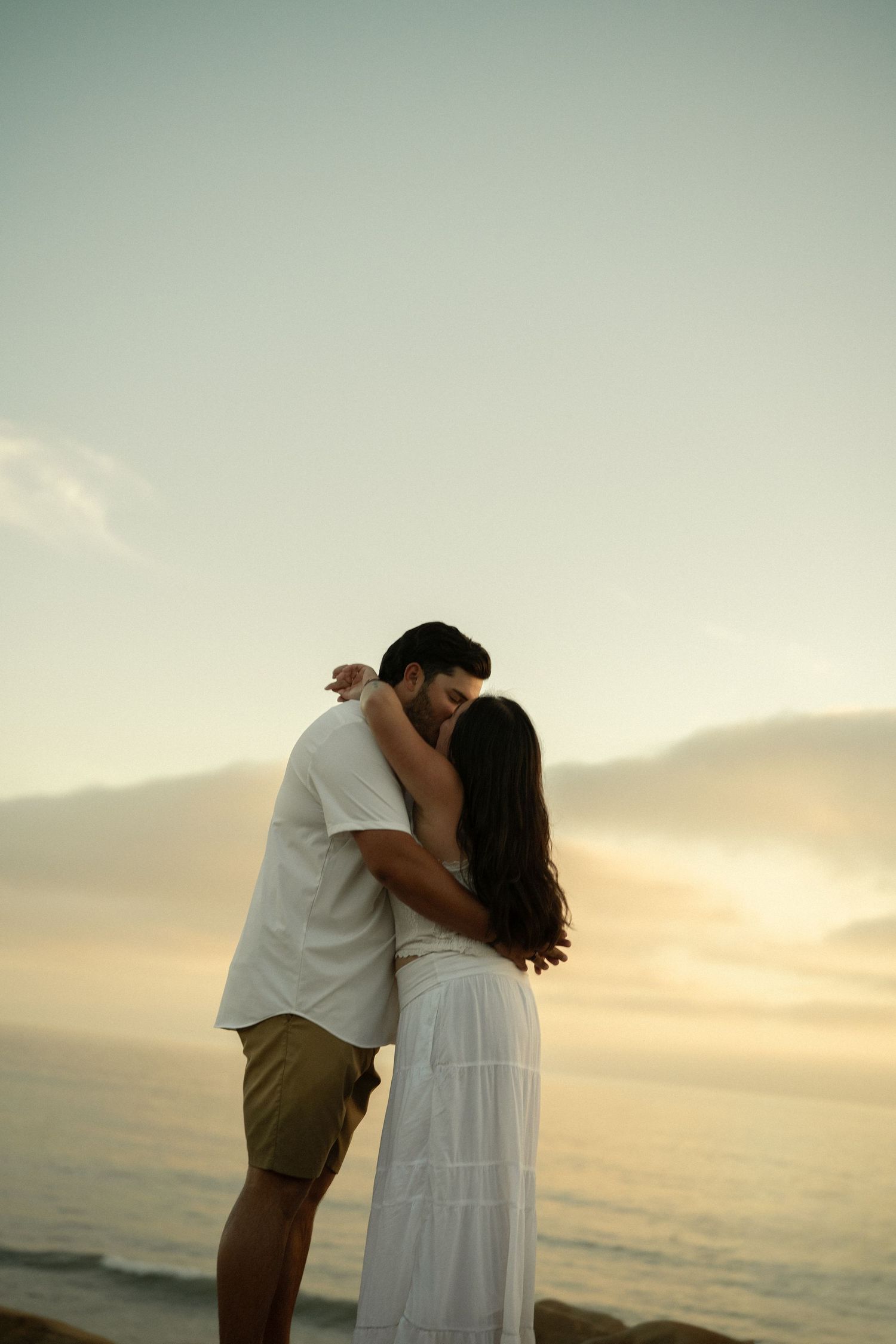 Couple sharing tender kiss at beach during sunset, woman in white dress embracing man in white shirt.