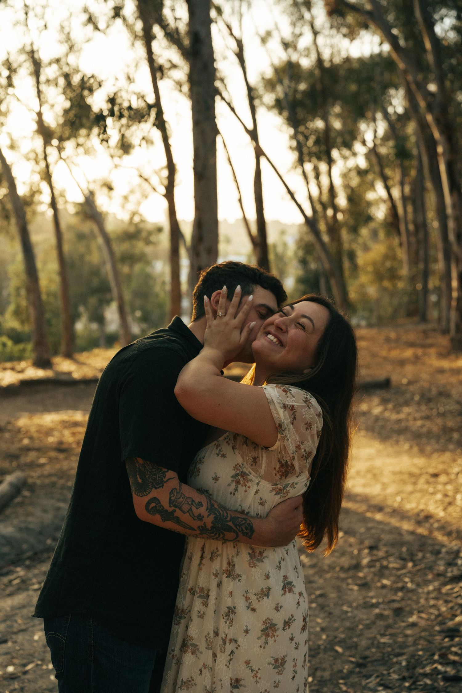 Woman laughing joyfully as her partner embraces her from behind in a golden sunlit forest.