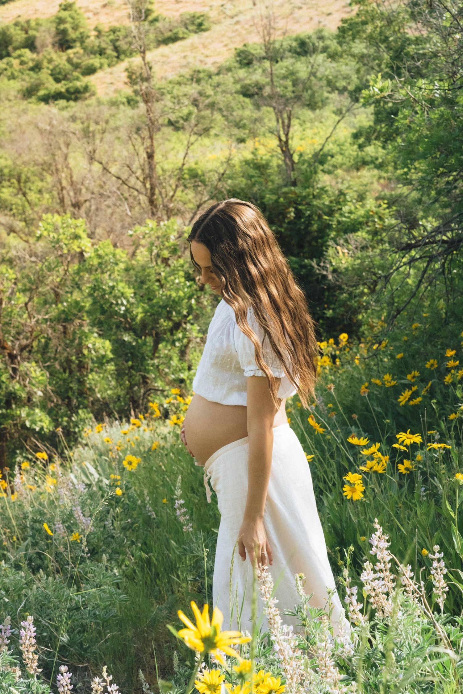 Pregnant woman in white dress standing among wildflowers and green hills, looking down at her baby bump.