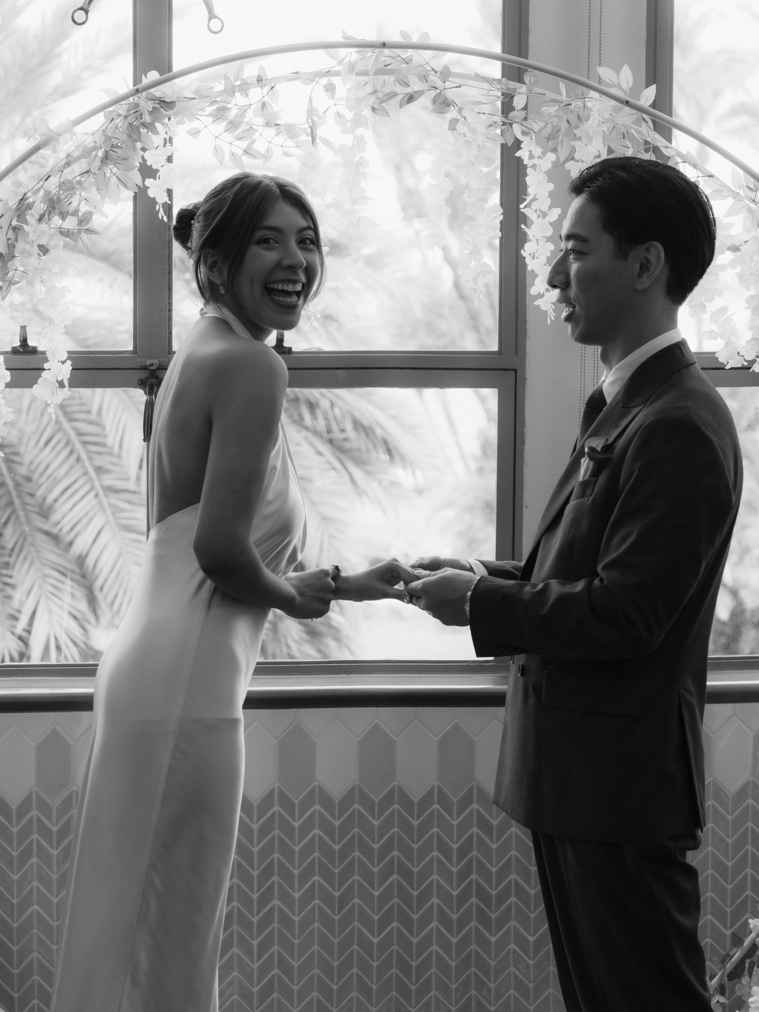 Black and white wedding photo of a laughing bride in a sleek gown holding hands with groom under a floral arch.