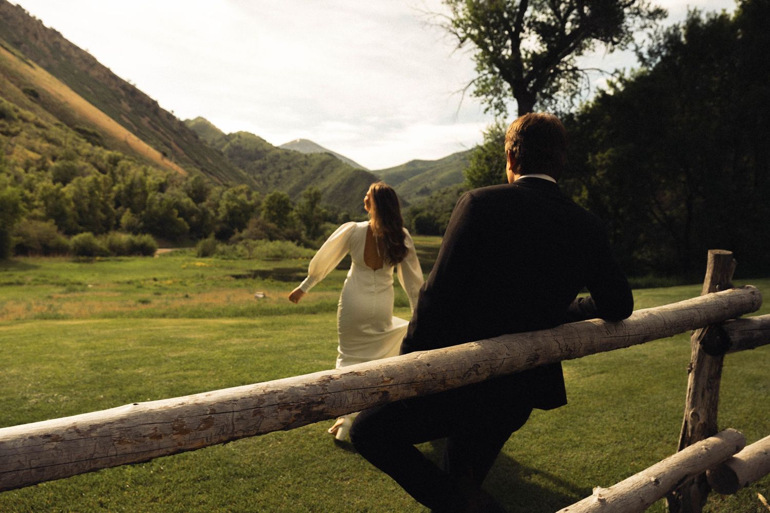 Two people share a quiet moment on a wooden fence in a serene mountain setting.