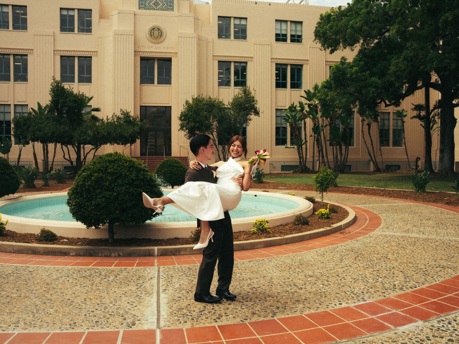 Groom carrying bride in white dress by ornate fountain in courtyard of elegant building on wedding day.
