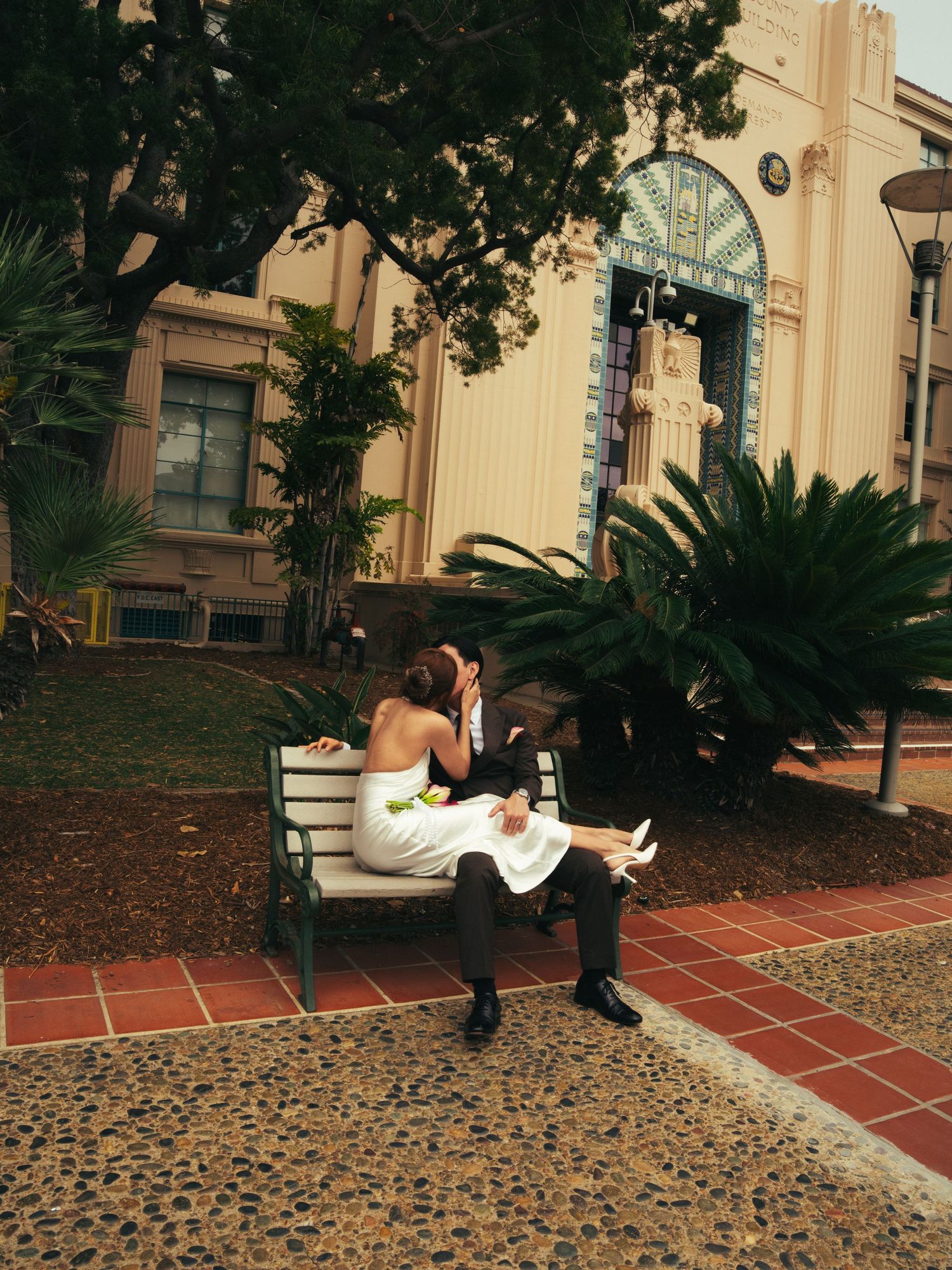 A bride and groom sit closely on a bench outside a historic building surrounded by palm trees.