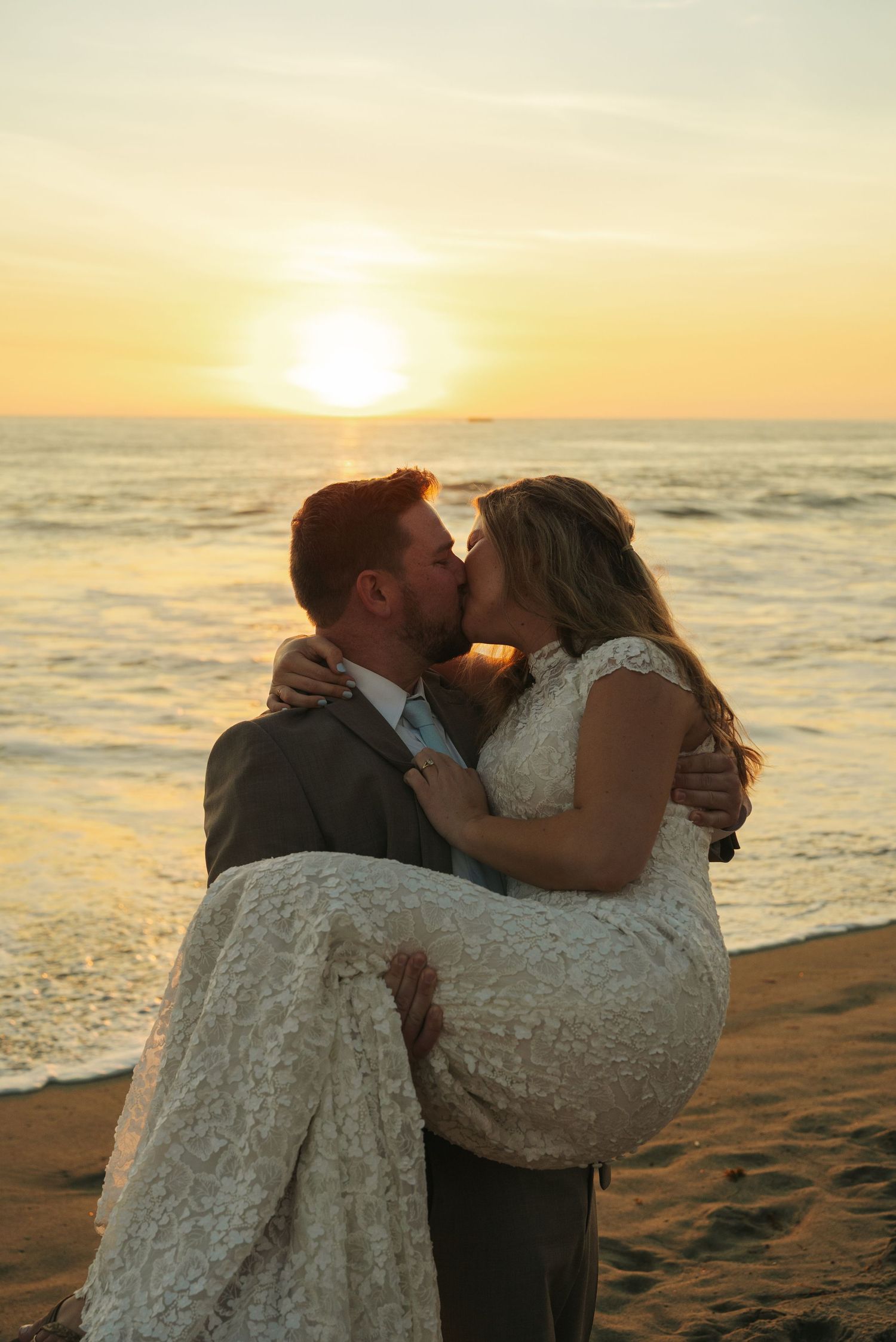 Groom carrying bride in his arms sharing a kiss on the beach during a golden sunset.