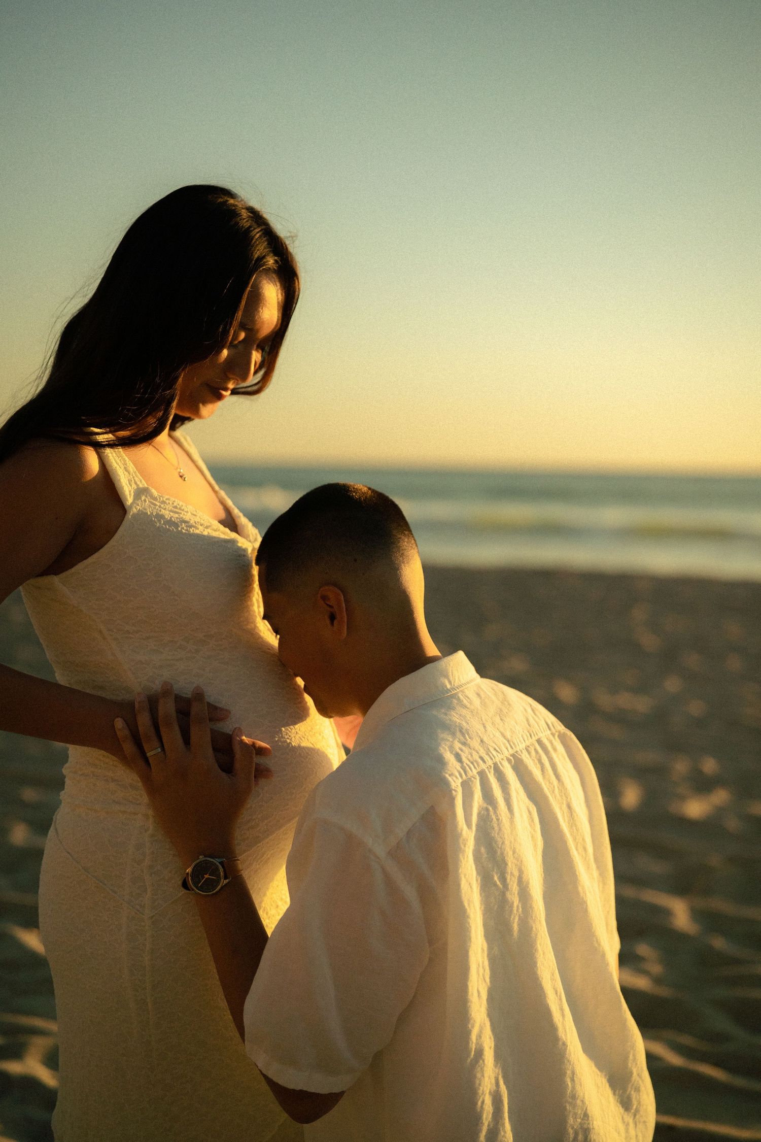Man tenderly kissing pregnant woman's belly on sandy beach during golden hour sunset.