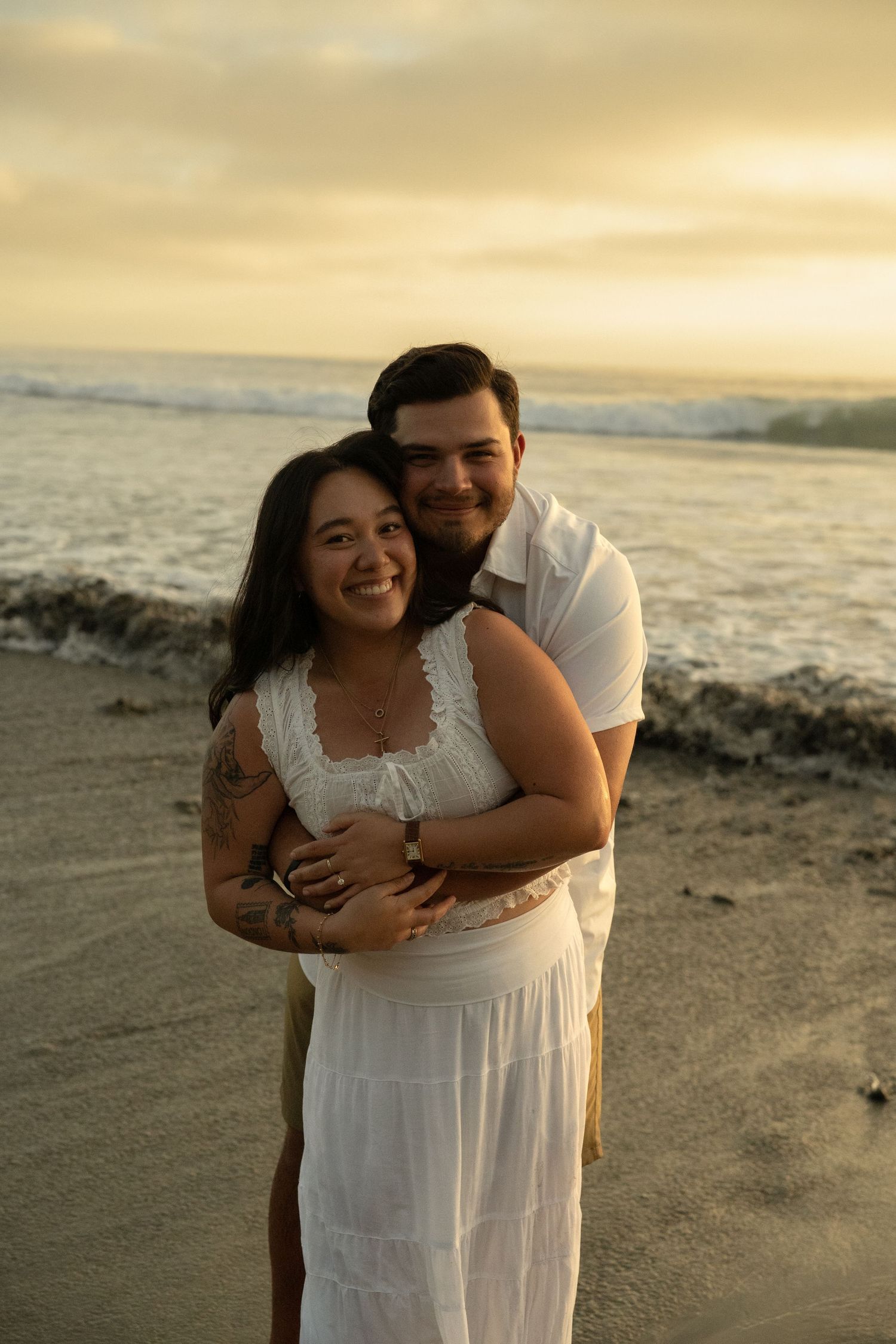 Couple embracing on beach at golden sunset, woman in white dress smiling, man in white shirt hugging her from behind.