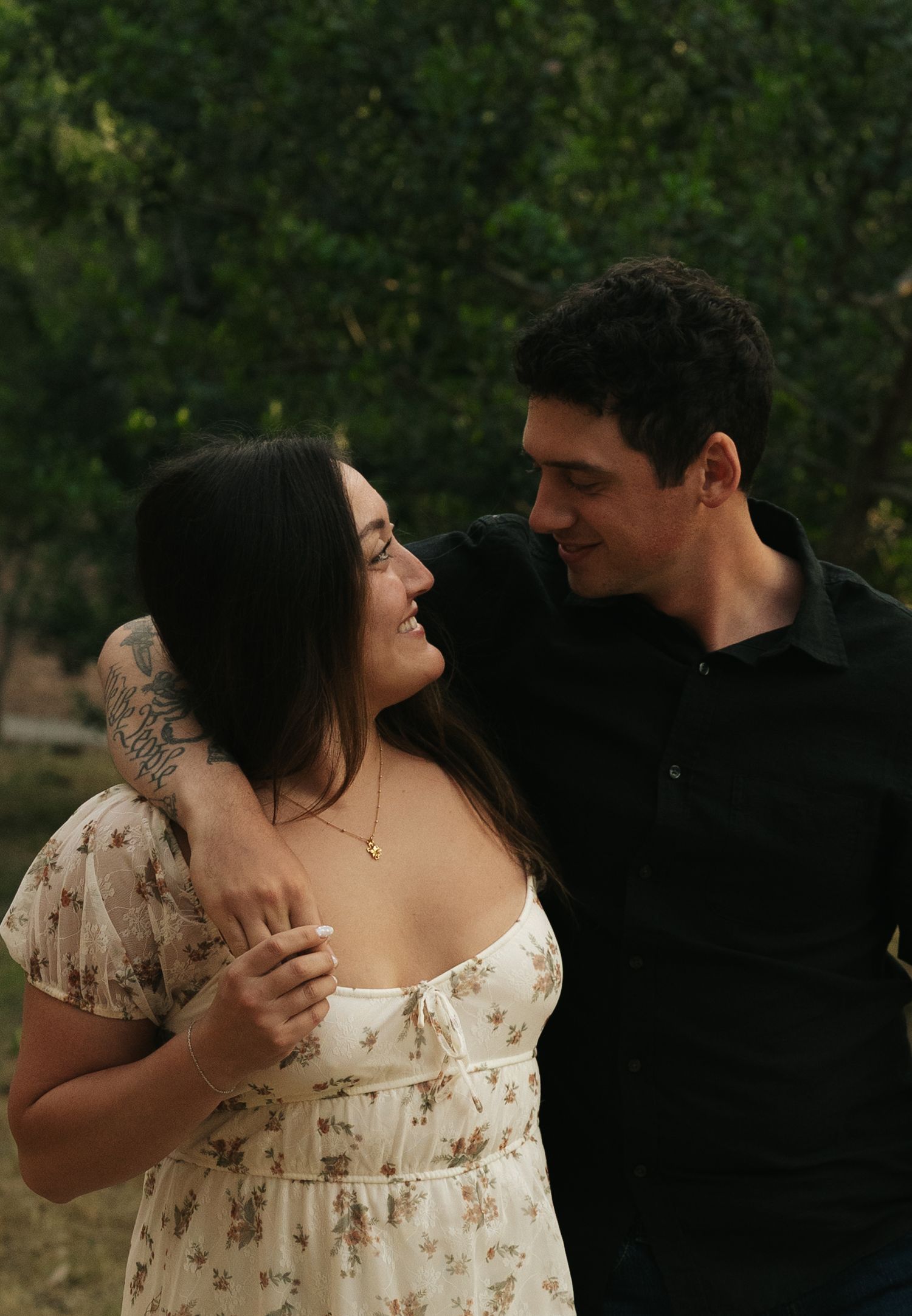 A smiling couple embraces outdoors, the woman in a floral dress and the man in a black shirt, sharing a tender moment.