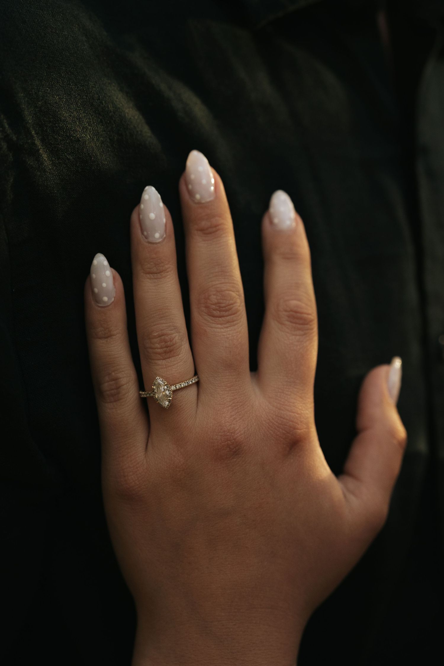 Close-up of a hand with white polka dot almond nails wearing a delicate diamond engagement ring.